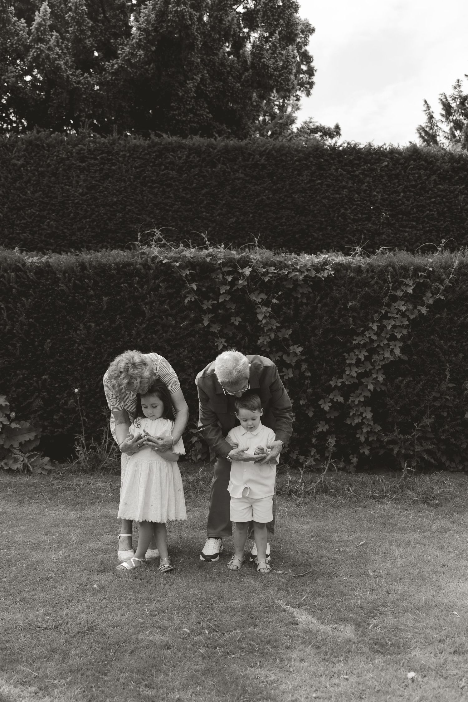 Black and white photo of elderly man bending over two young children standing on grass in front of tall hedge.