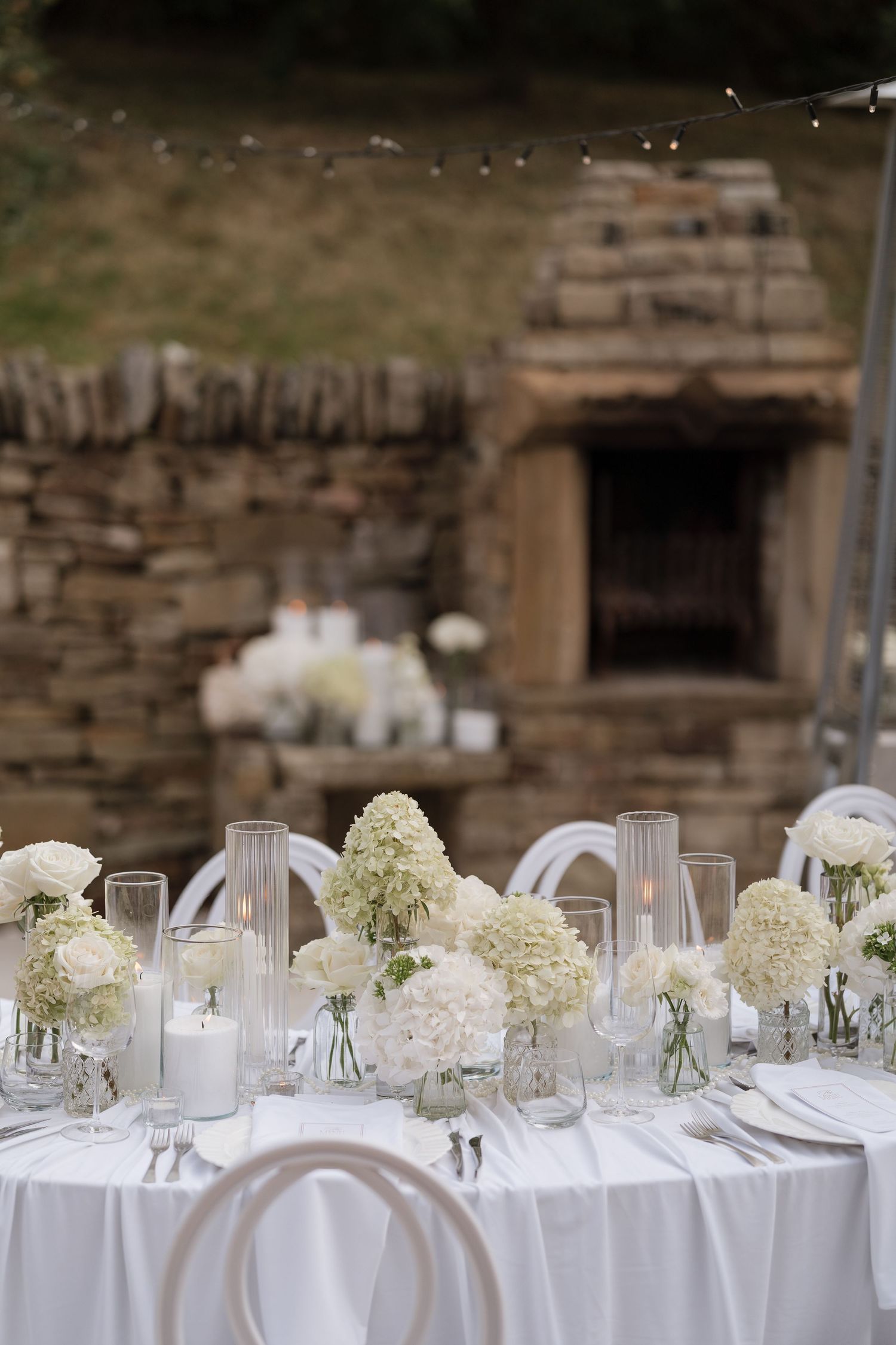 Elegant outdoor wedding reception table with white hydrangeas, candles, and glassware against a rustic stone wall backdrop.