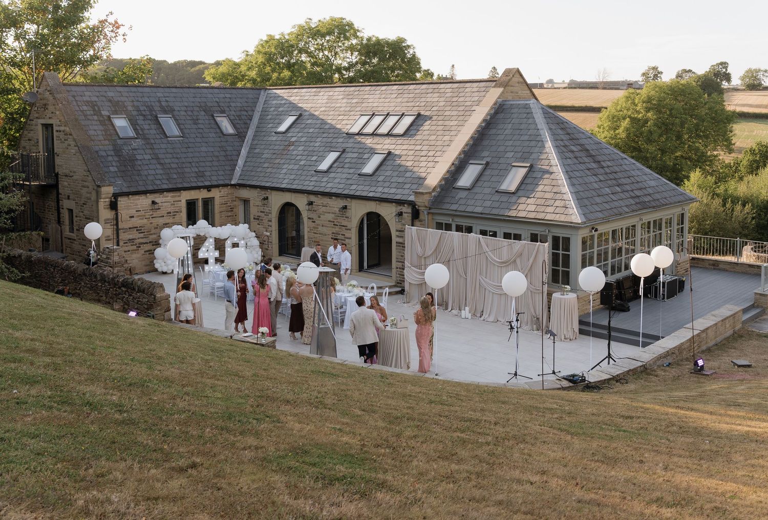 Stone barn wedding venue with guests gathered outside on patio, balloon decor, surrounded by green countryside at golden hour.