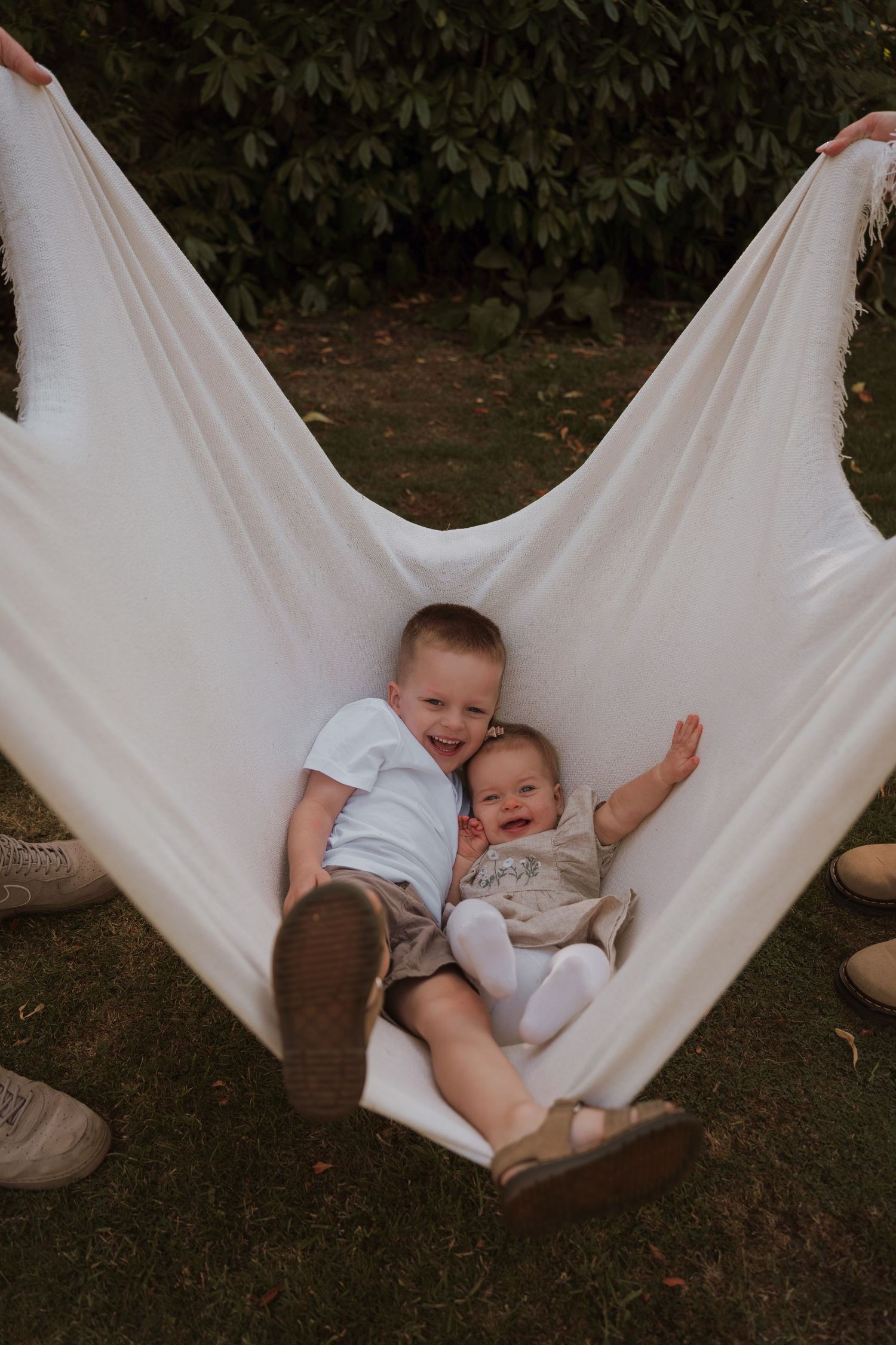 Two young children laughing and cuddling together in a white fabric hammock outdoors surrounded by lush green foliage.