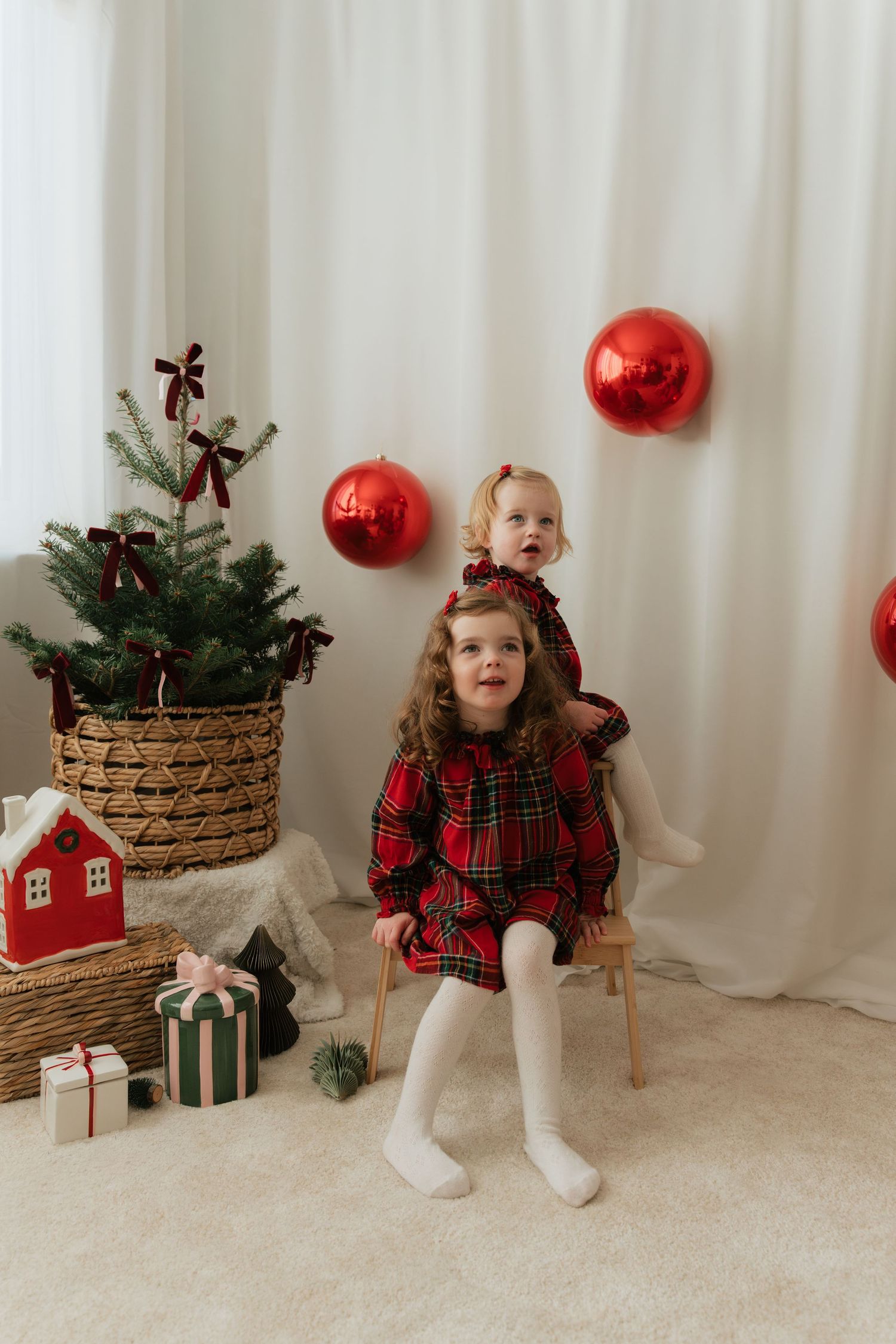 Two young girls in red plaid dresses pose near a decorated Christmas tree with gifts and red ornament balloons.