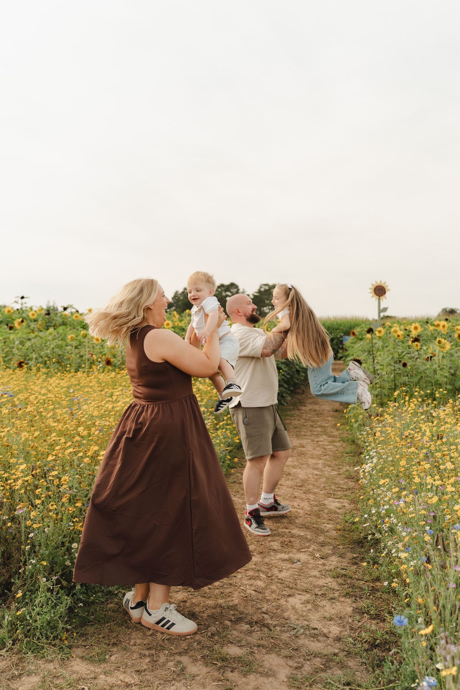 Family enjoying a sunny day in a sunflower field, with parents playfully lifting children on a dirt path.