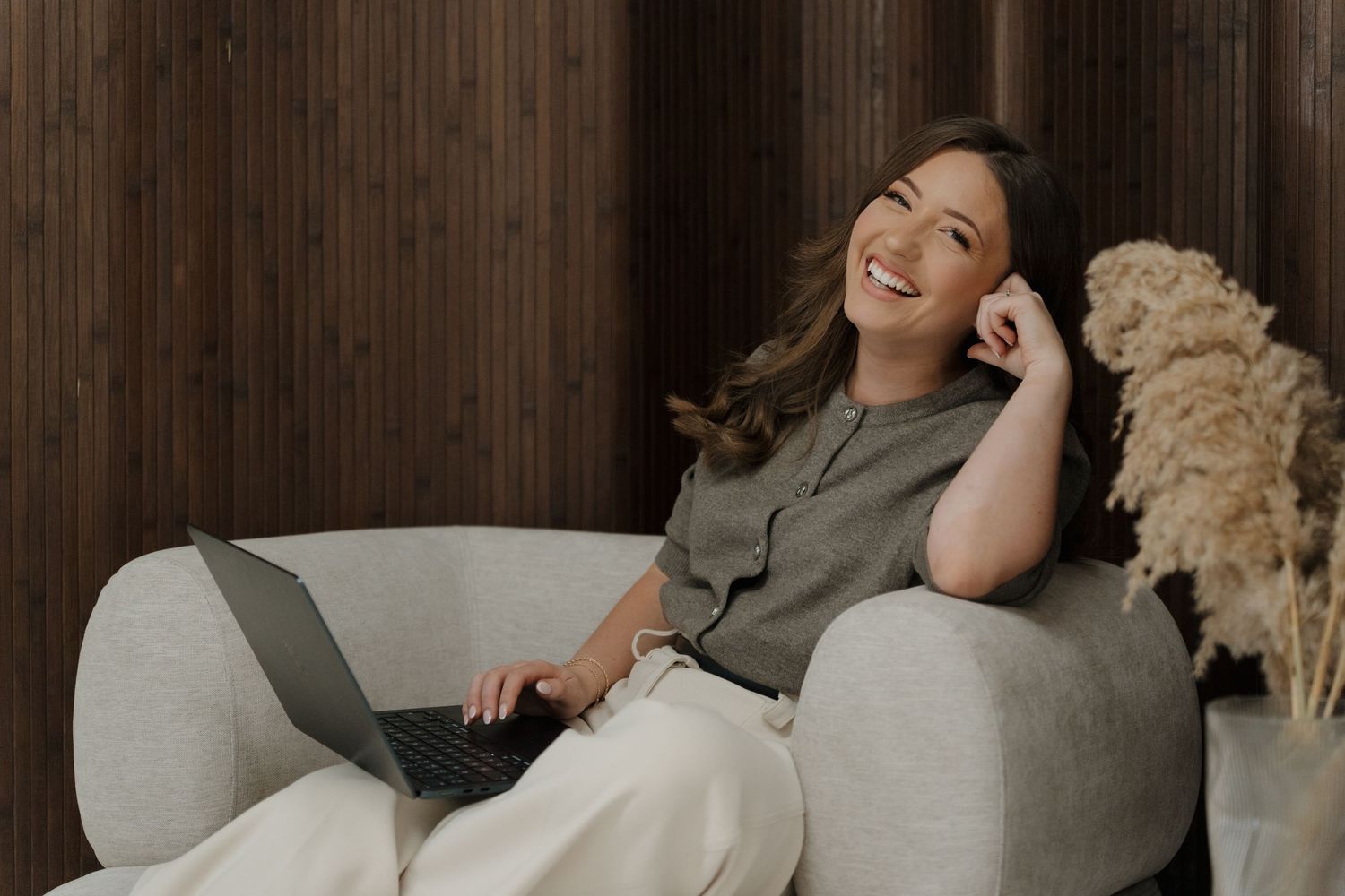 Brand photography Derby. Smiling woman sitting on a cozy sofa with a laptop, relaxing in a modern interior with warm wood paneling.