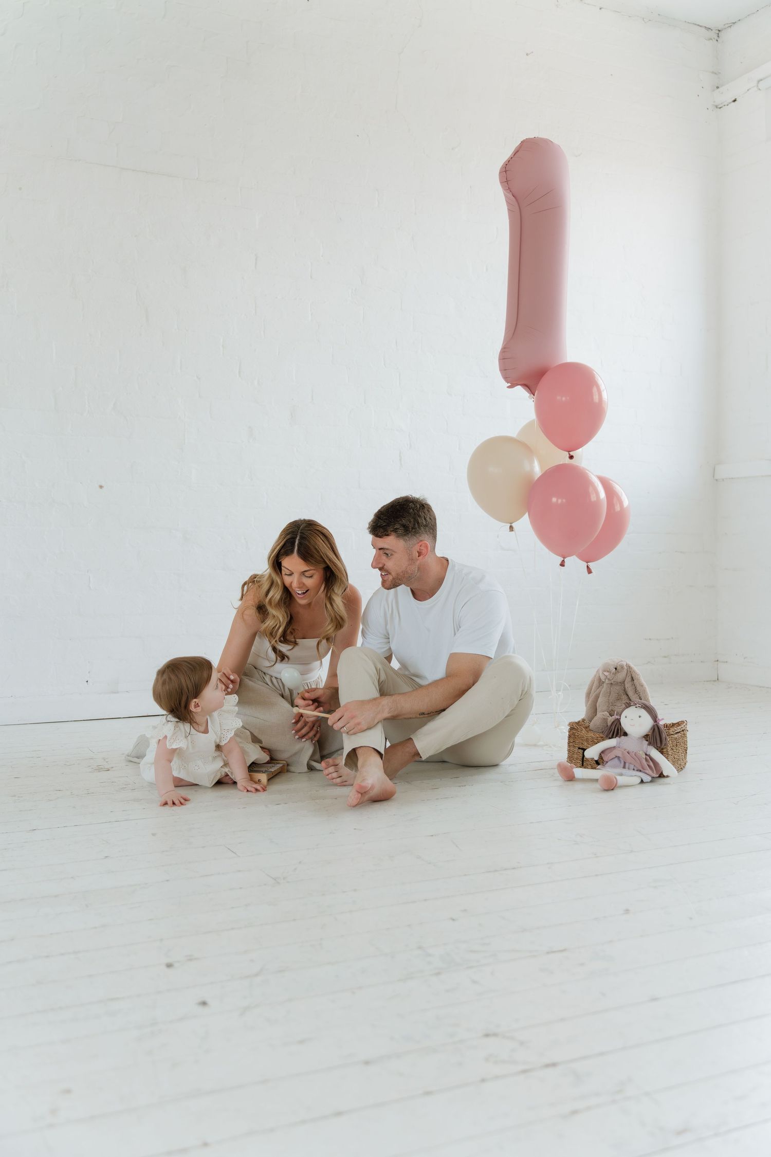 A young family celebrates their baby's first birthday with pink balloons and toys in a bright white room.