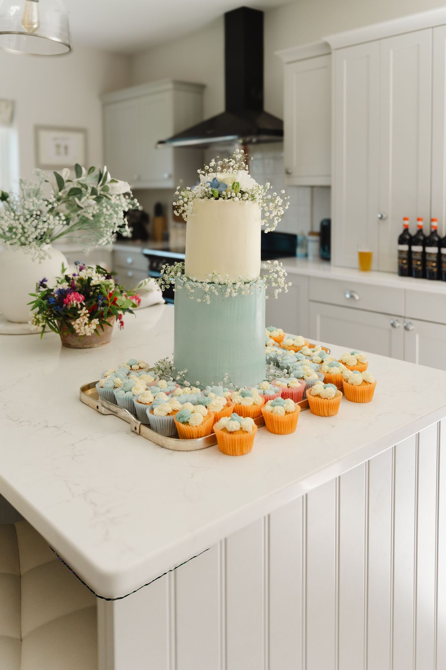 Small pumpkins and floral decorations surround a pale blue wedding cake on a white marble kitchen counter.
