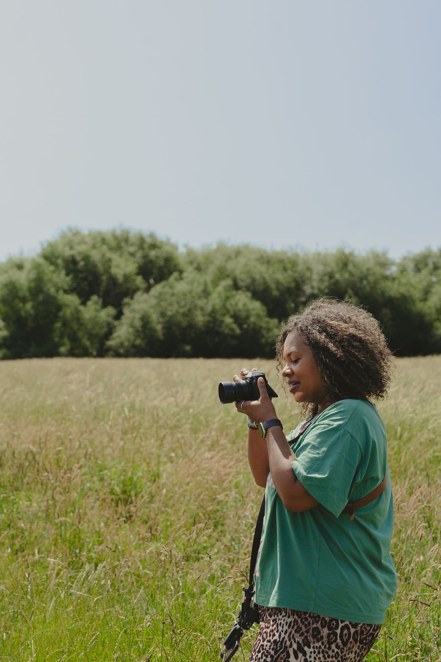 A photographer in a green shirt takes pictures in a sunny meadow with trees in the background.