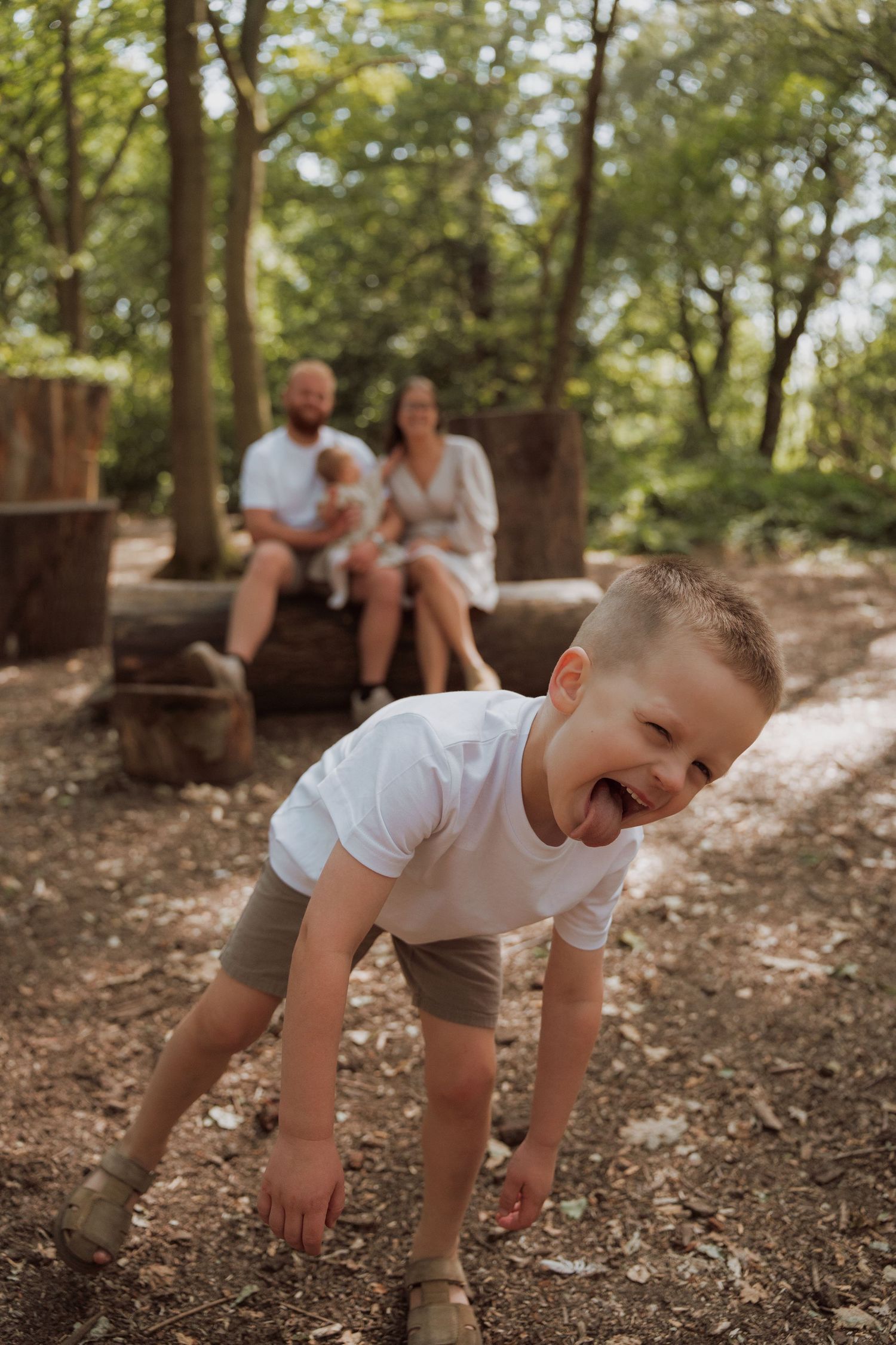 A laughing boy plays in a forest while his parents sit on a log with a baby in the background.