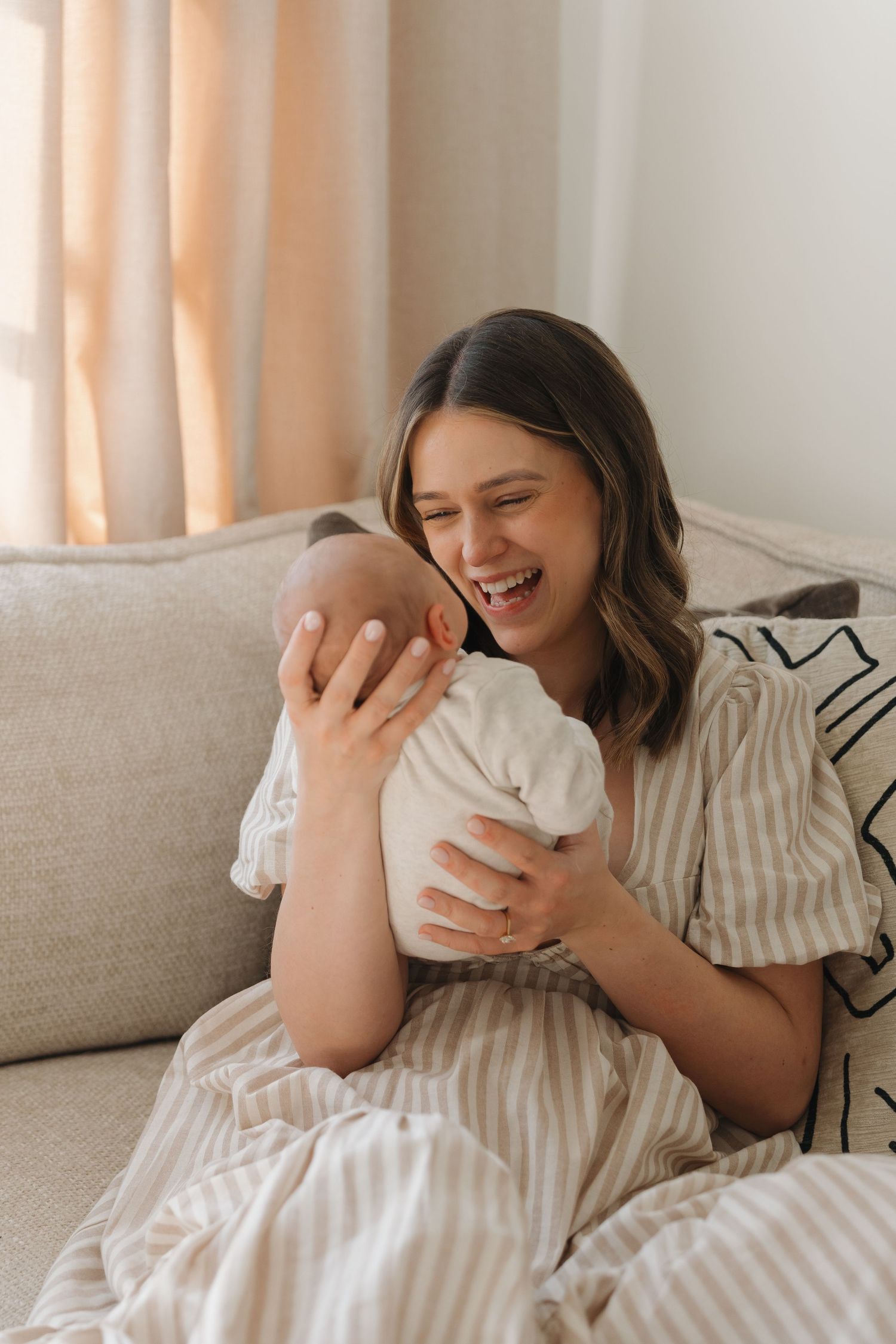 Happy young mother laughing while holding her newborn baby on a cozy beige sofa at home. Home Newborn Photography Derbyshire Shots by Maz.