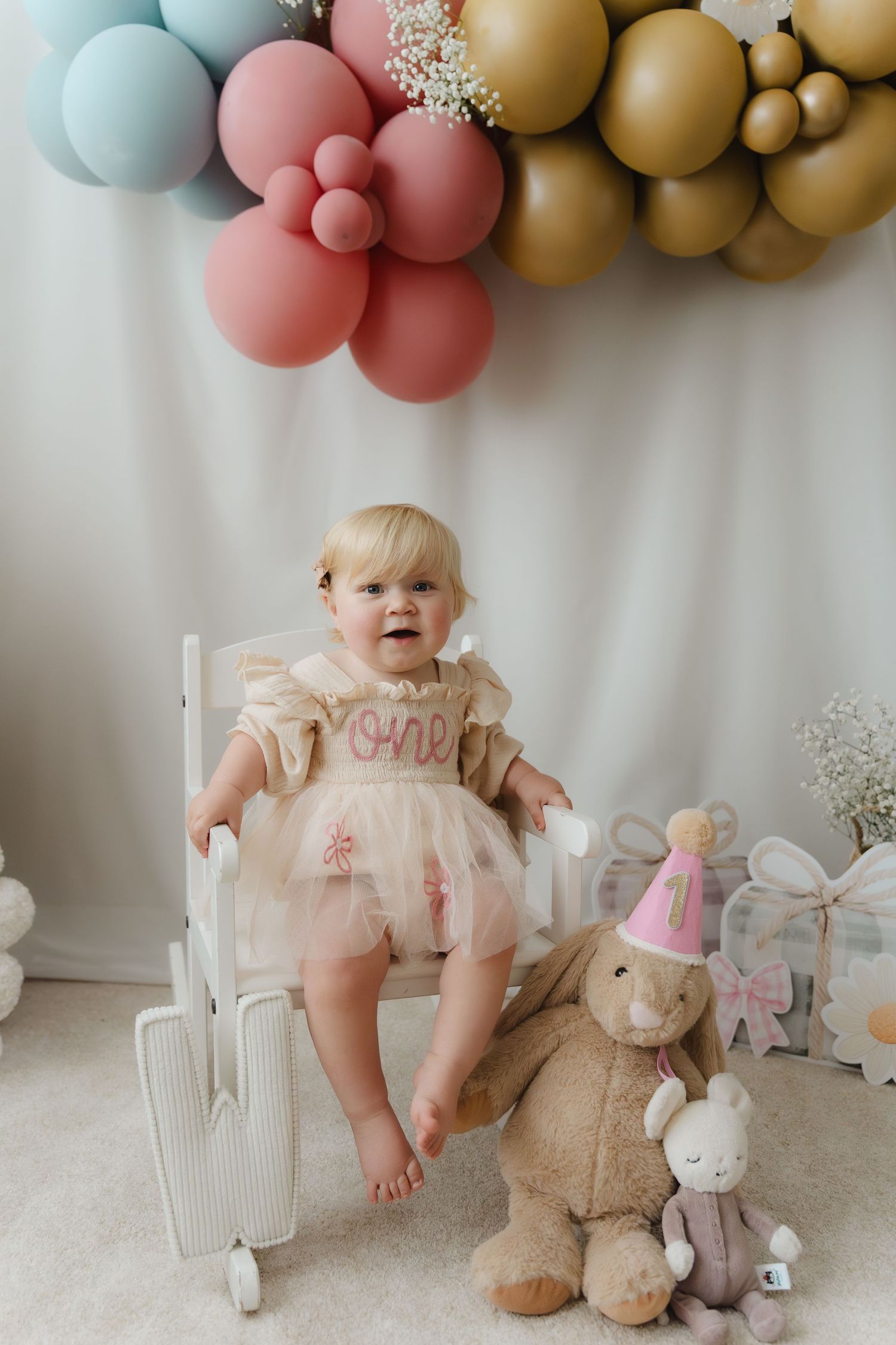 A toddler in a pink dress sits on a white chair under decorative balloons with a plush bunny toy nearby.