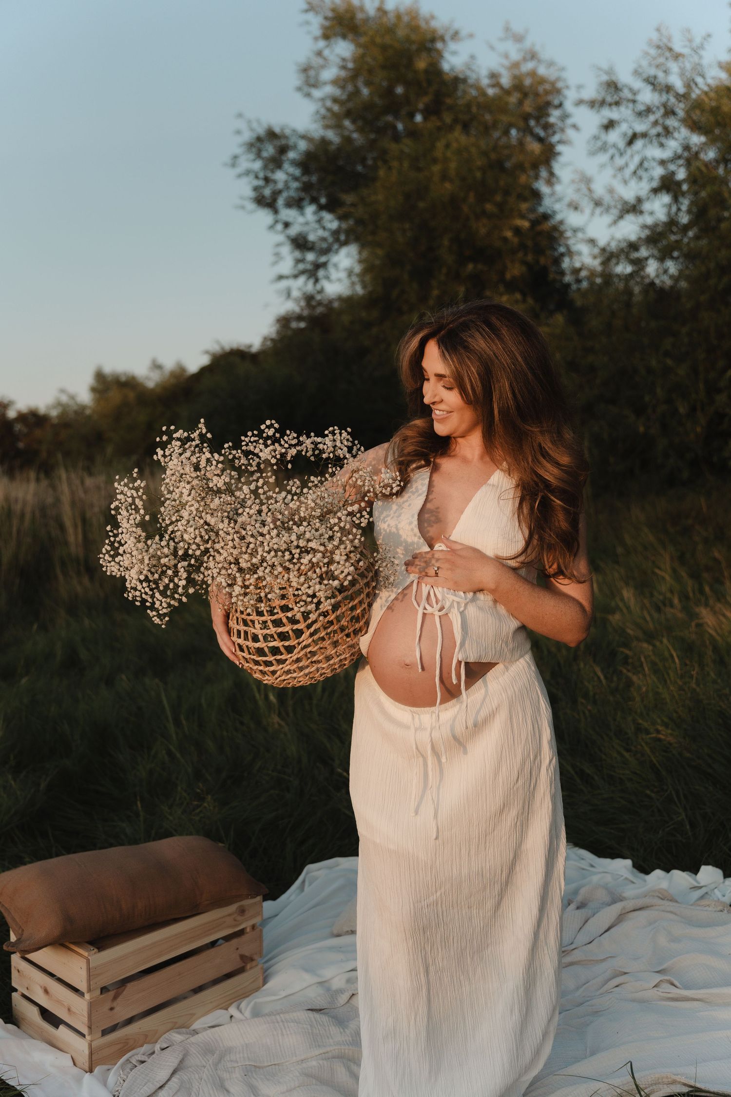 Woman in white dress sits on blanket outdoors holding dried floral arrangements in vintage wooden crates at sunset.