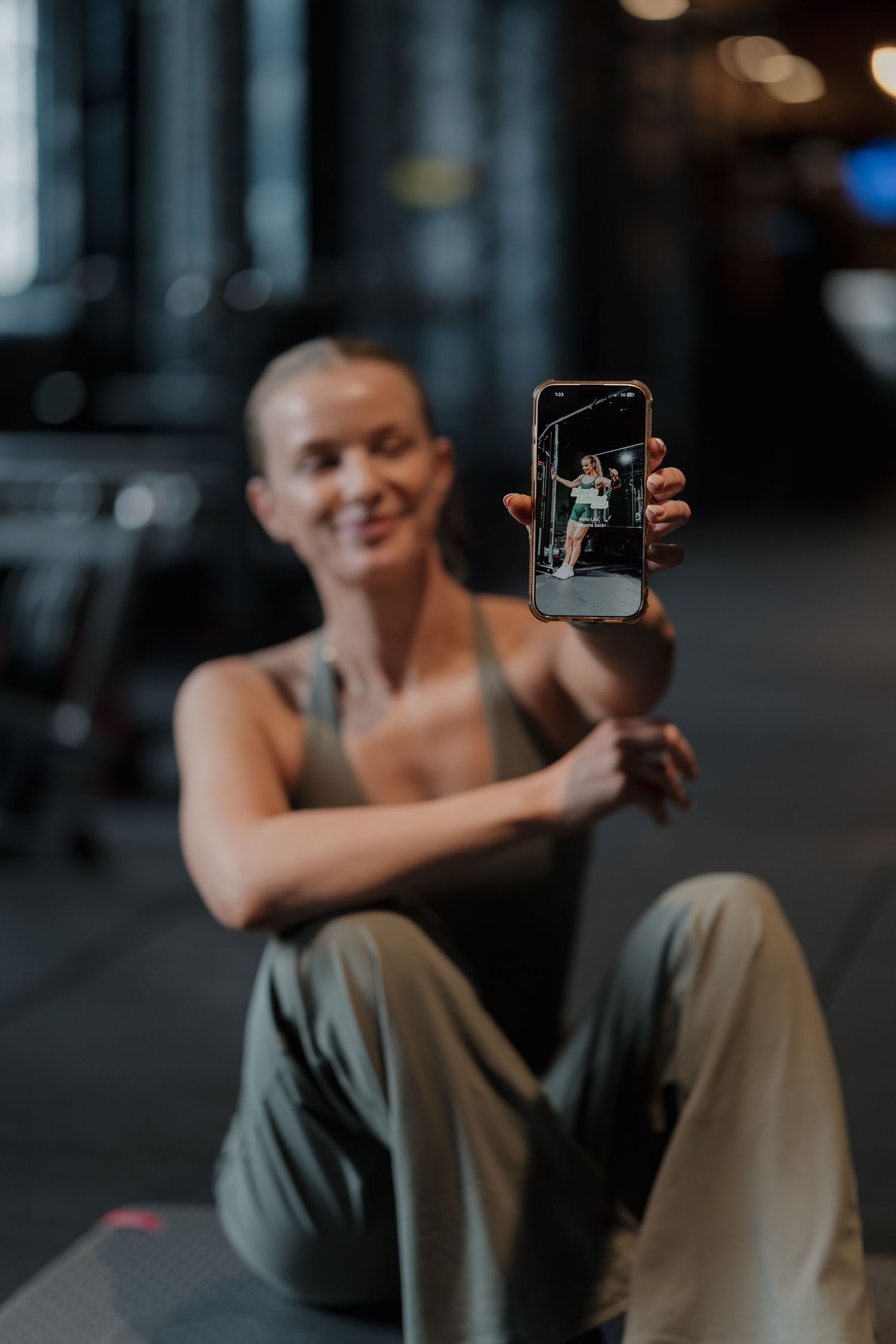 Smiling fit woman in gym sitting on floor holding up smartphone showing workout video on screen. Brand photography Derbyshire Shots by Maz