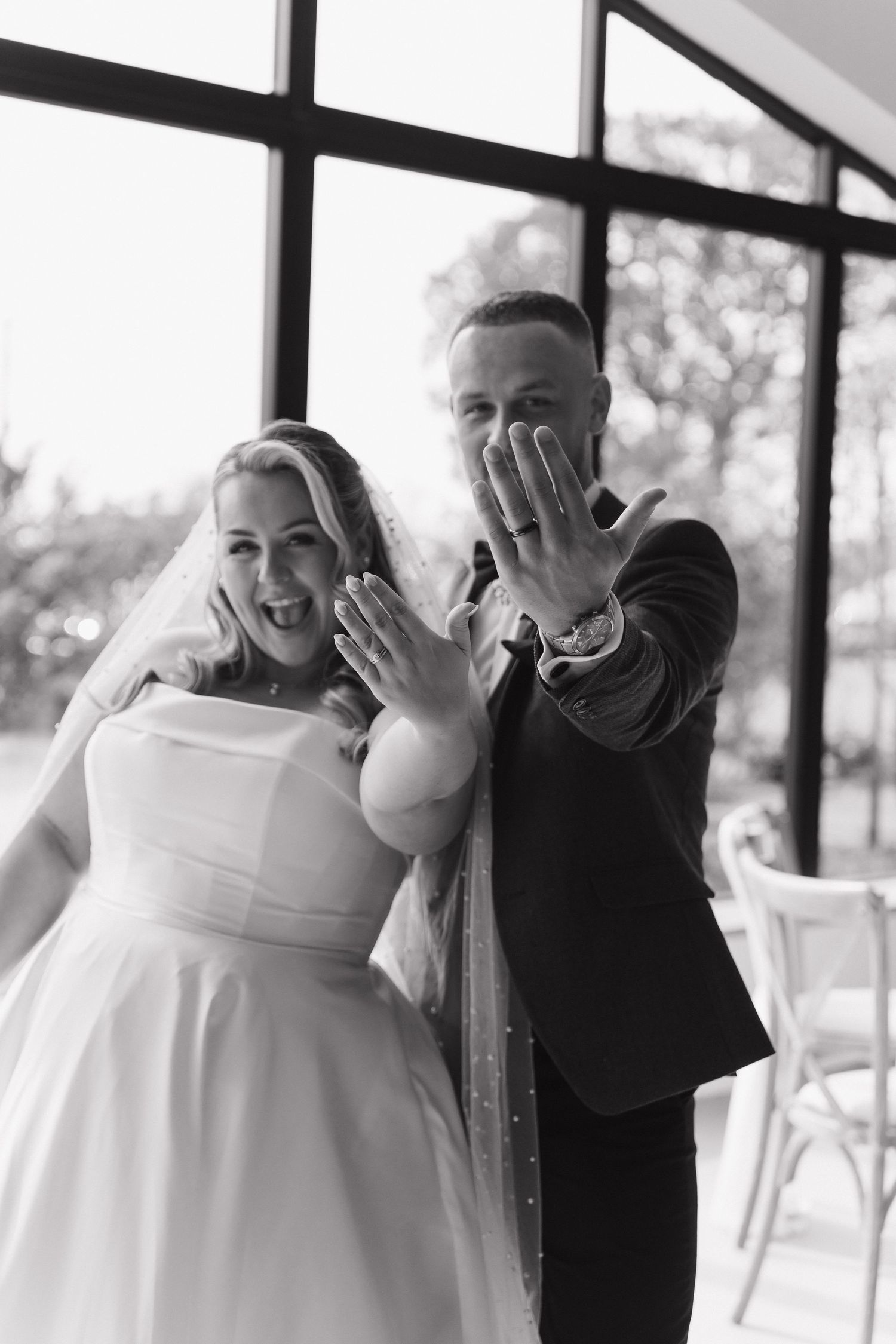 Black and white photo of newlyweds showing off wedding rings, bride in white gown and veil, groom in dark suit.