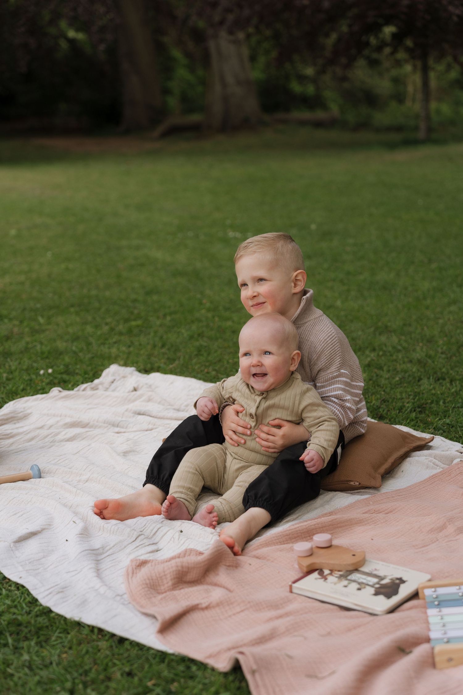 Two young siblings sitting together on a blanket in a green park, older child hugging baby, both smiling happily outdoors.