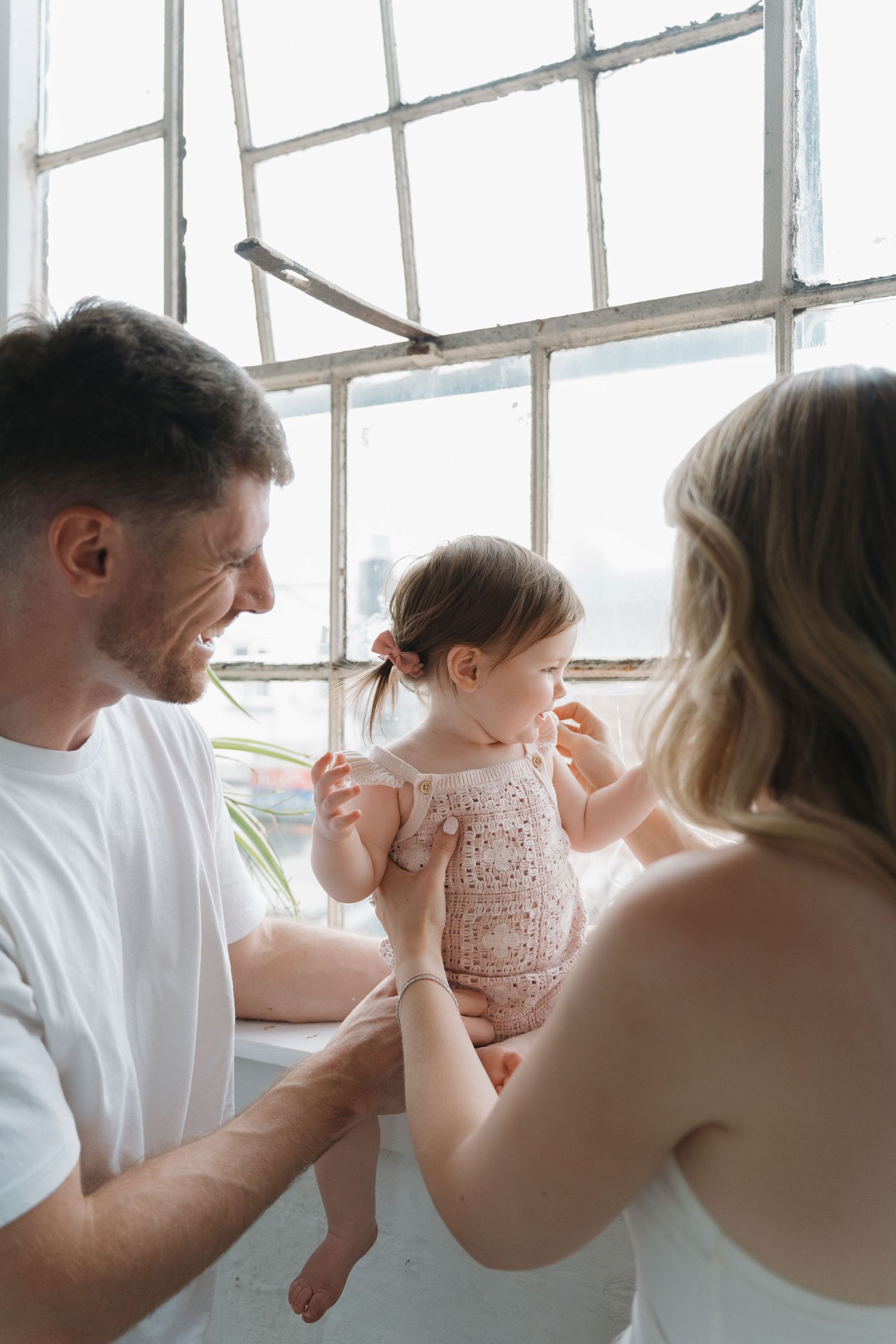 Happy parents smiling and playing with their baby girl in a bright, sunlit room near large windows.