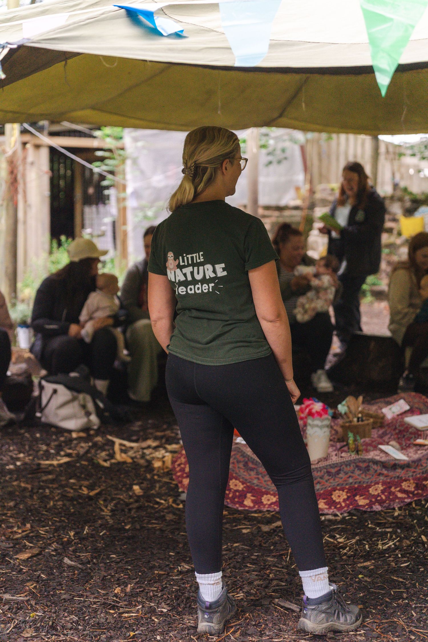 Person wearing a dark t-shirt and leggings stands at an outdoor event under a canopy with scattered rugs on the ground.