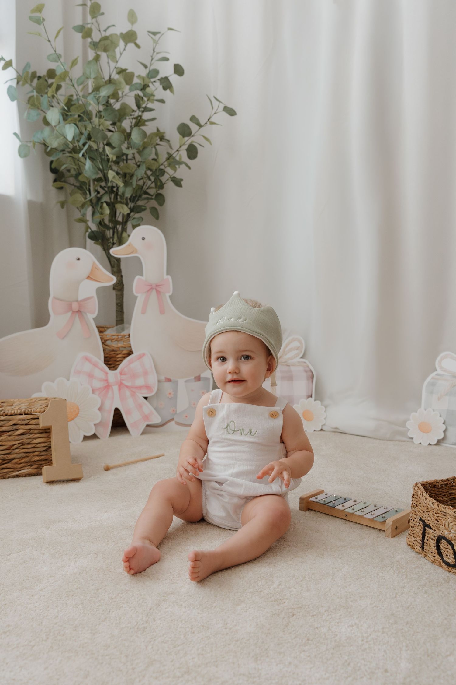 Baby in white outfit sits on plush carpet with decorative ducks and floral arrangements in light, airy nursery setting.