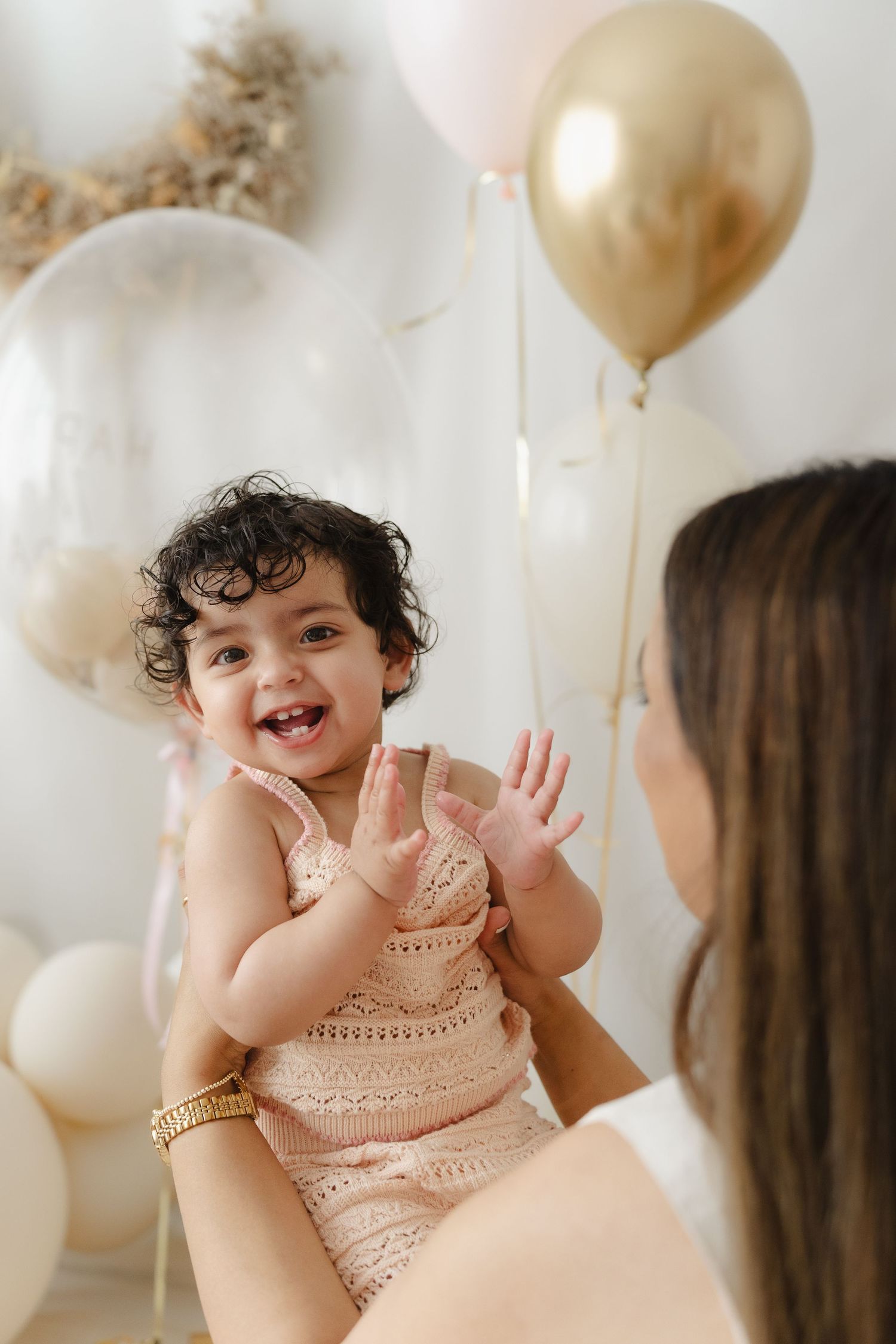 A baby in a pink lace dress laughs joyfully while surrounded by gold and white balloons at a celebration.
