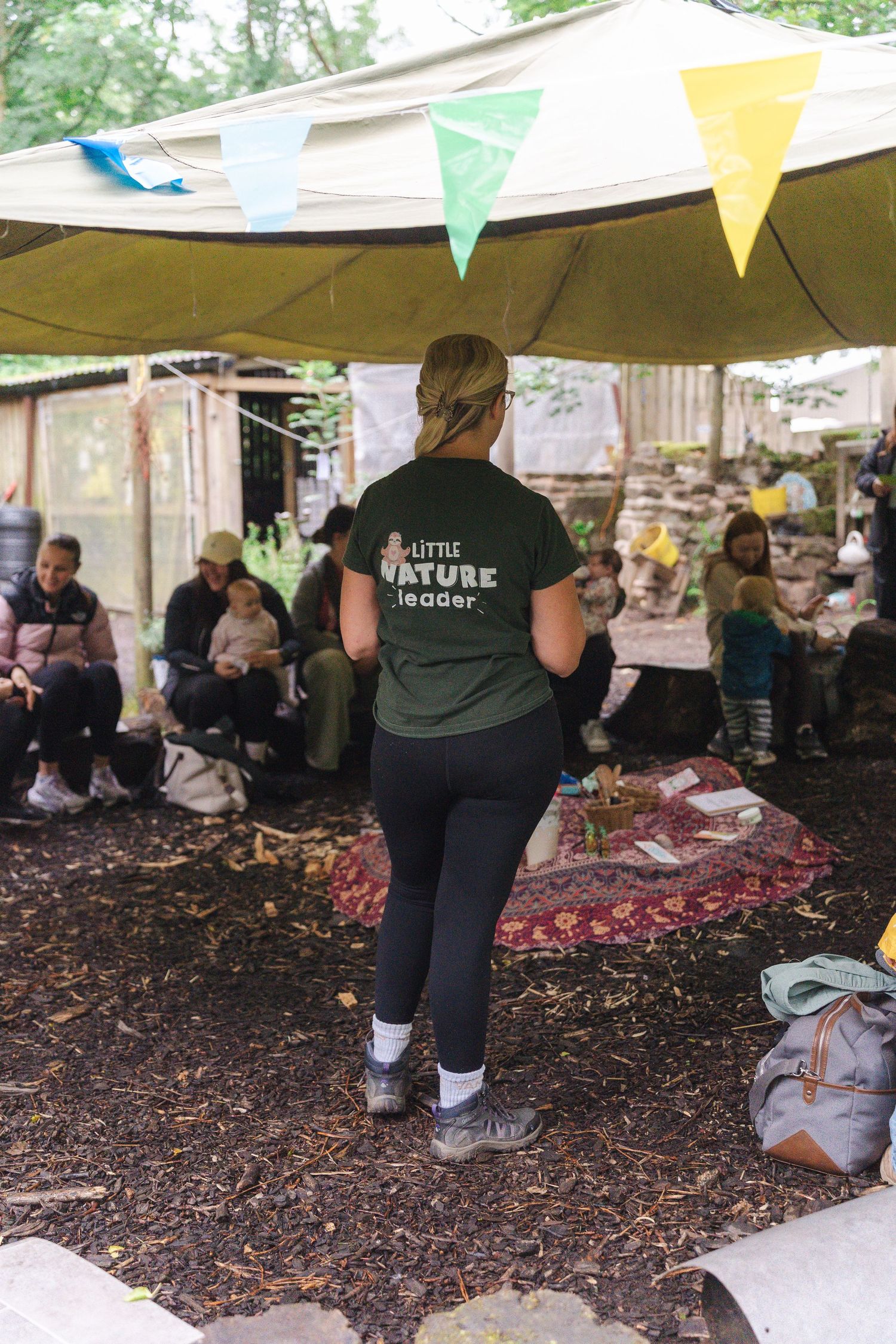 A Little Nature Leader instructor stands before a group of children and adults gathered under a colorful bunting canopy outdoors.