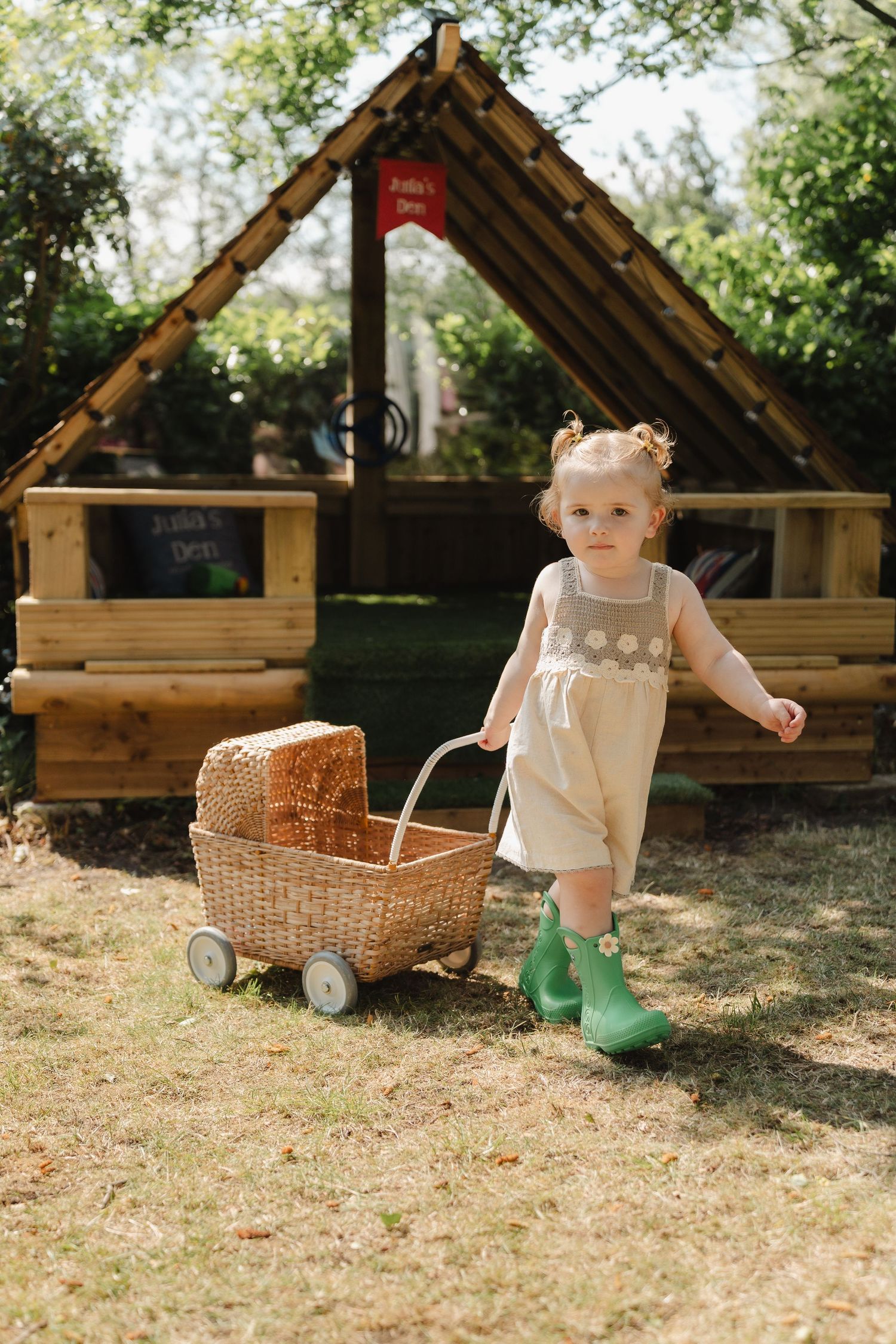 Toddler girl in green boots pushes wicker toy pram in sunny garden beside wooden playhouse.