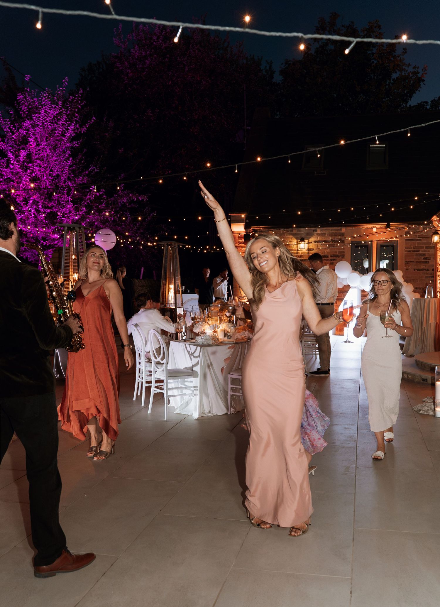Woman in pink gown dancing at outdoor wedding reception with string lights and guests celebrating at night.