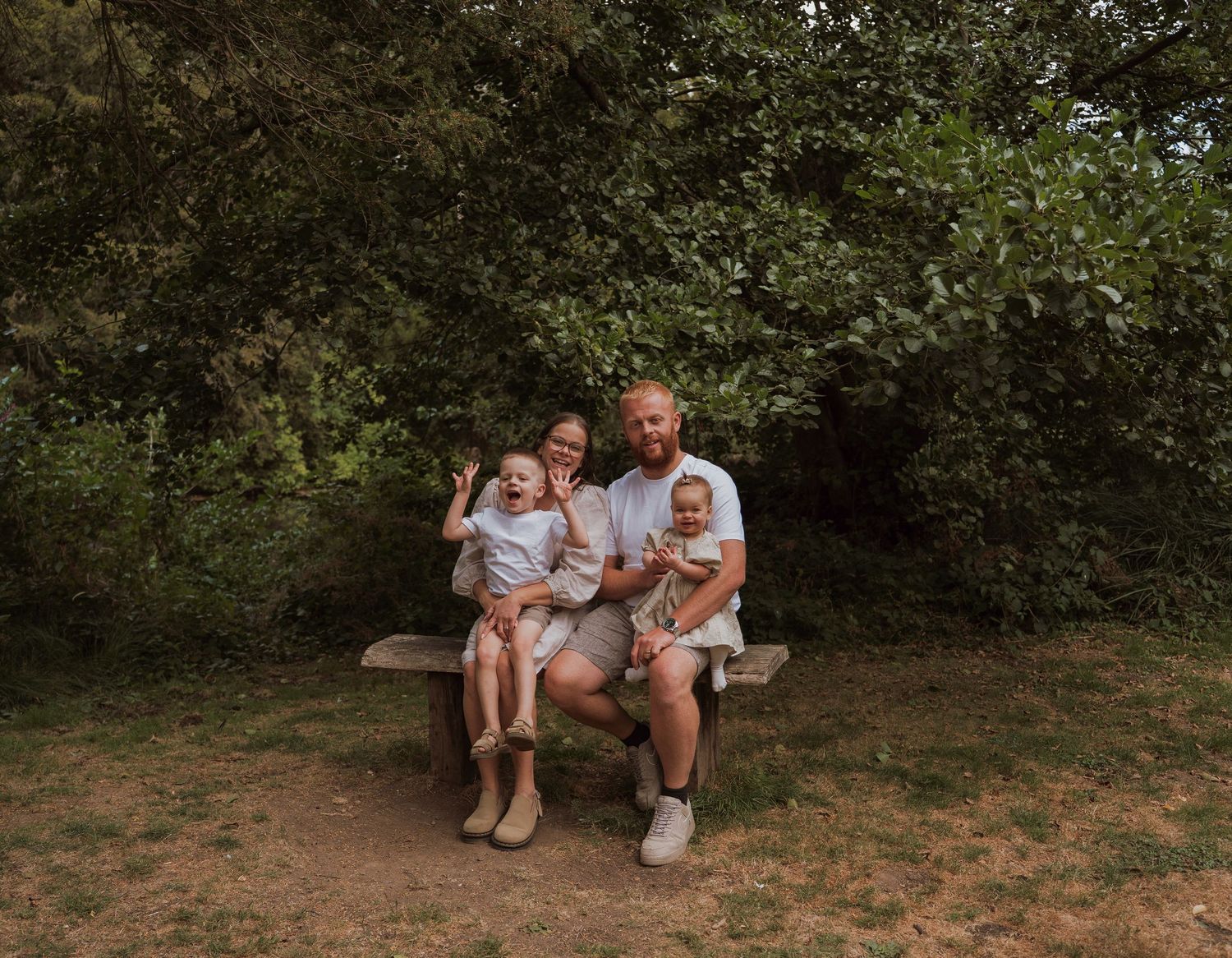 Family of four sitting on a wooden bench surrounded by lush green trees in a natural outdoor setting.