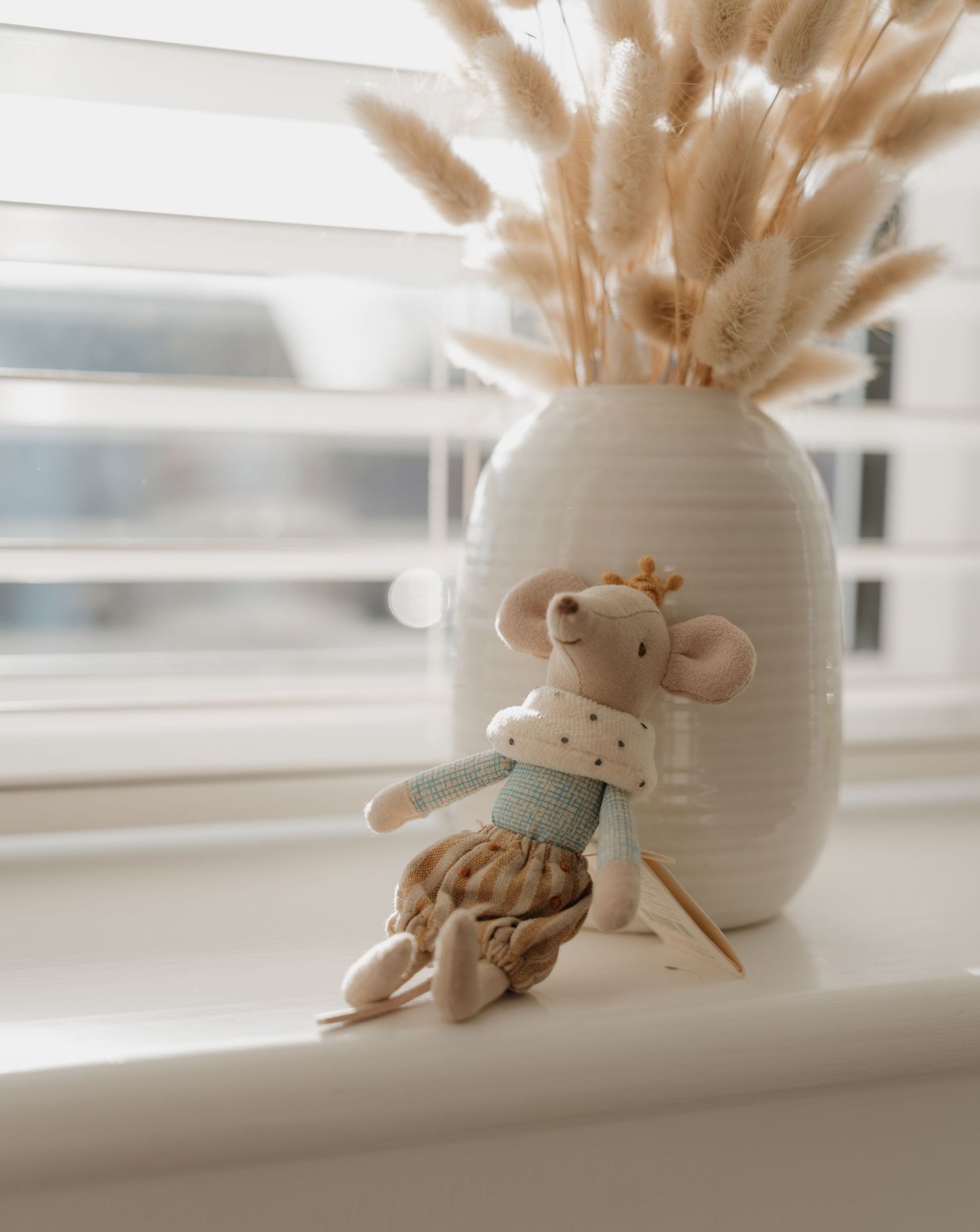 A small mouse toy figurine sits on a windowsill beside a white vase filled with dried pampas grass.