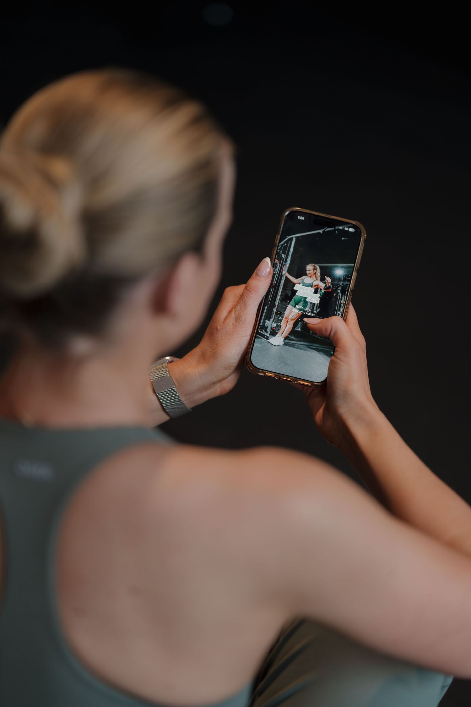 A blonde woman in a gray tank top films a fitness video on her smartphone in a dark gym setting.