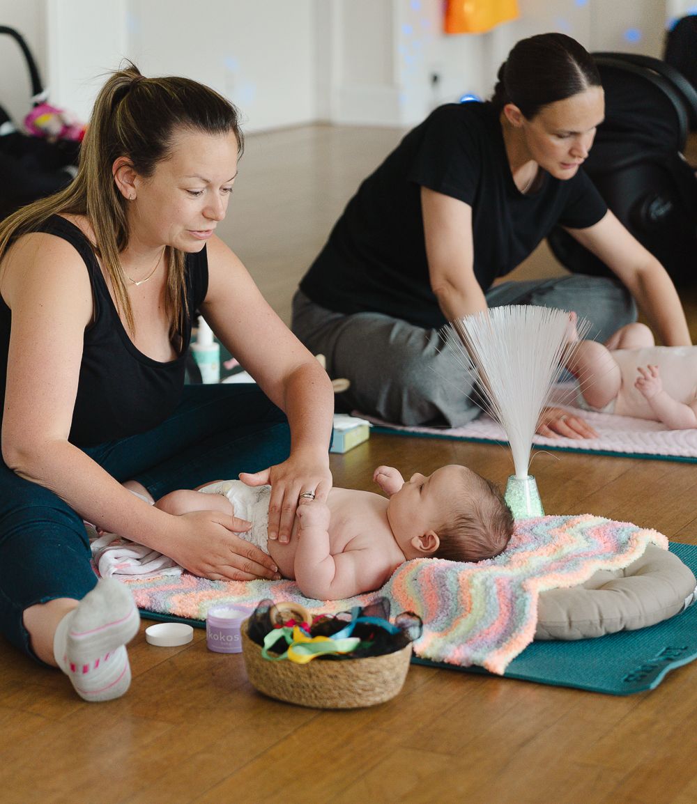 A baby receives a gentle massage during an infant massage class with several participants on yoga mats.