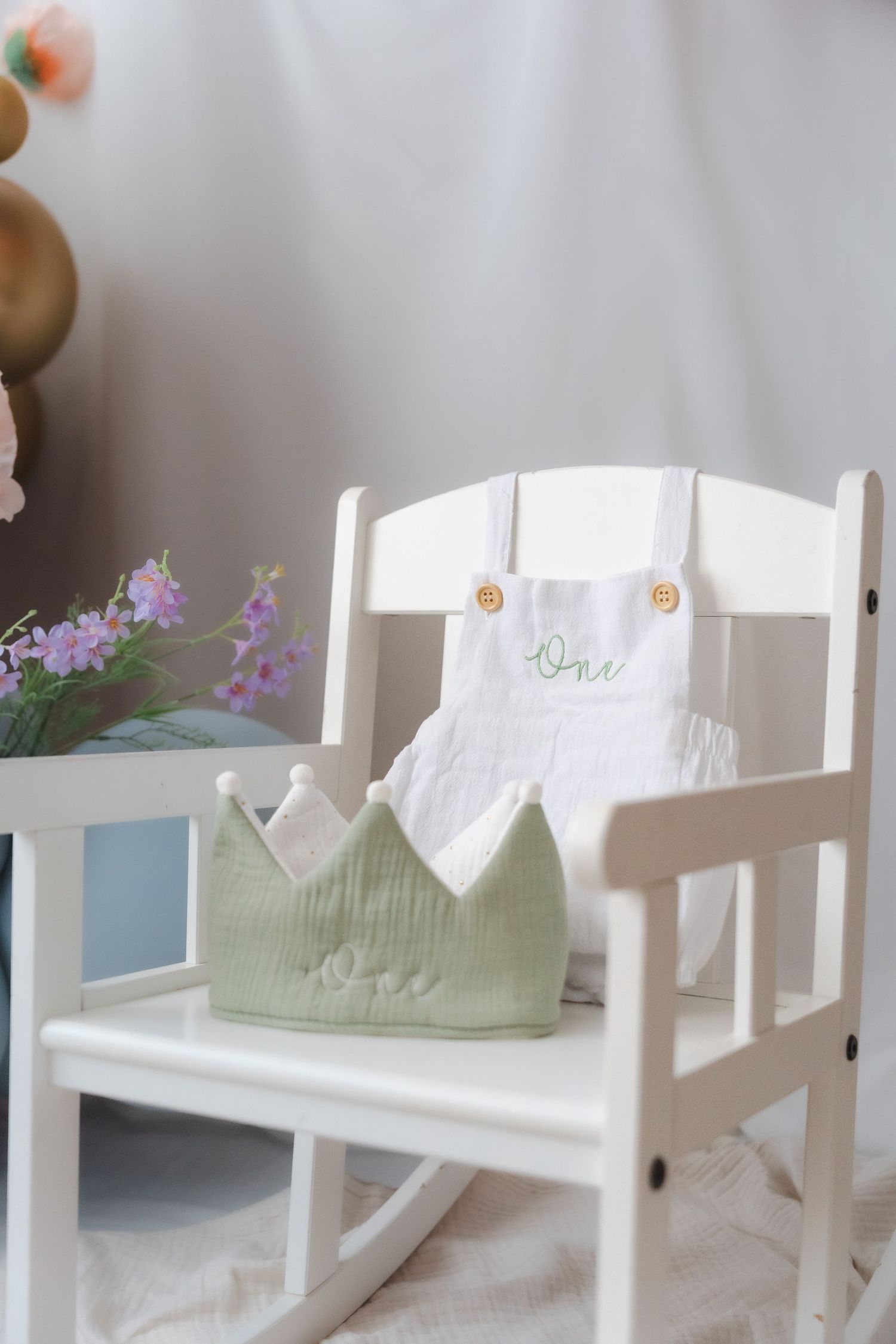 White wooden rocking chairs with soft green cushions sit in a row near a window with purple flowers in view.