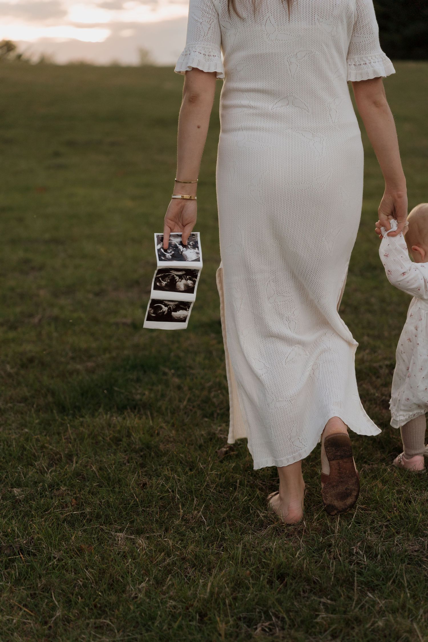 A person in a white dress holds a polaroid photograph while walking on grass.