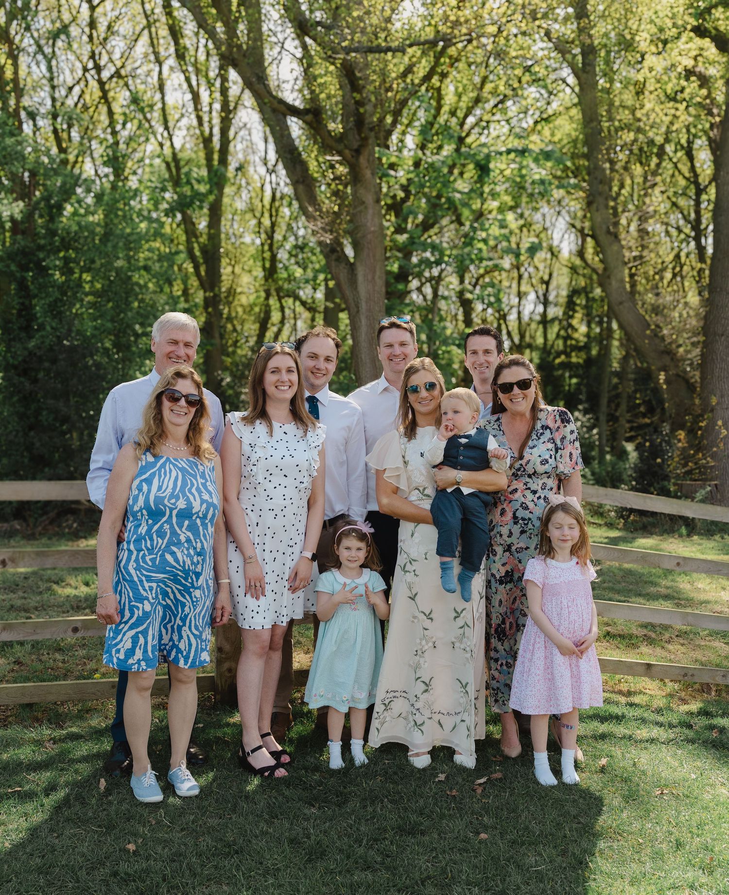 A group of people in light summer attire pose together on a grassy lawn with trees in the background.
