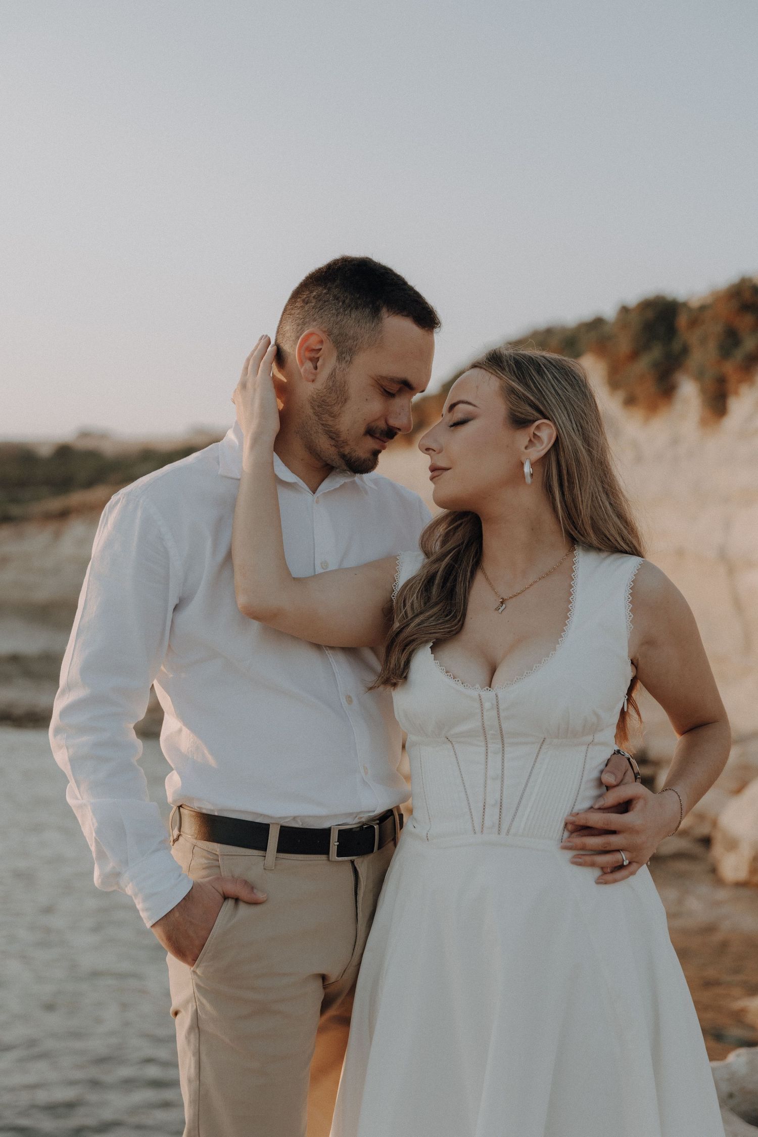 A couple in wedding attire share a romantic moment on a coastal cliff at sunset.