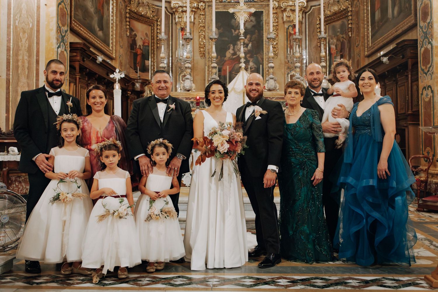 Large wedding party including flower girls and family members pose together in front of decorated church altar.
