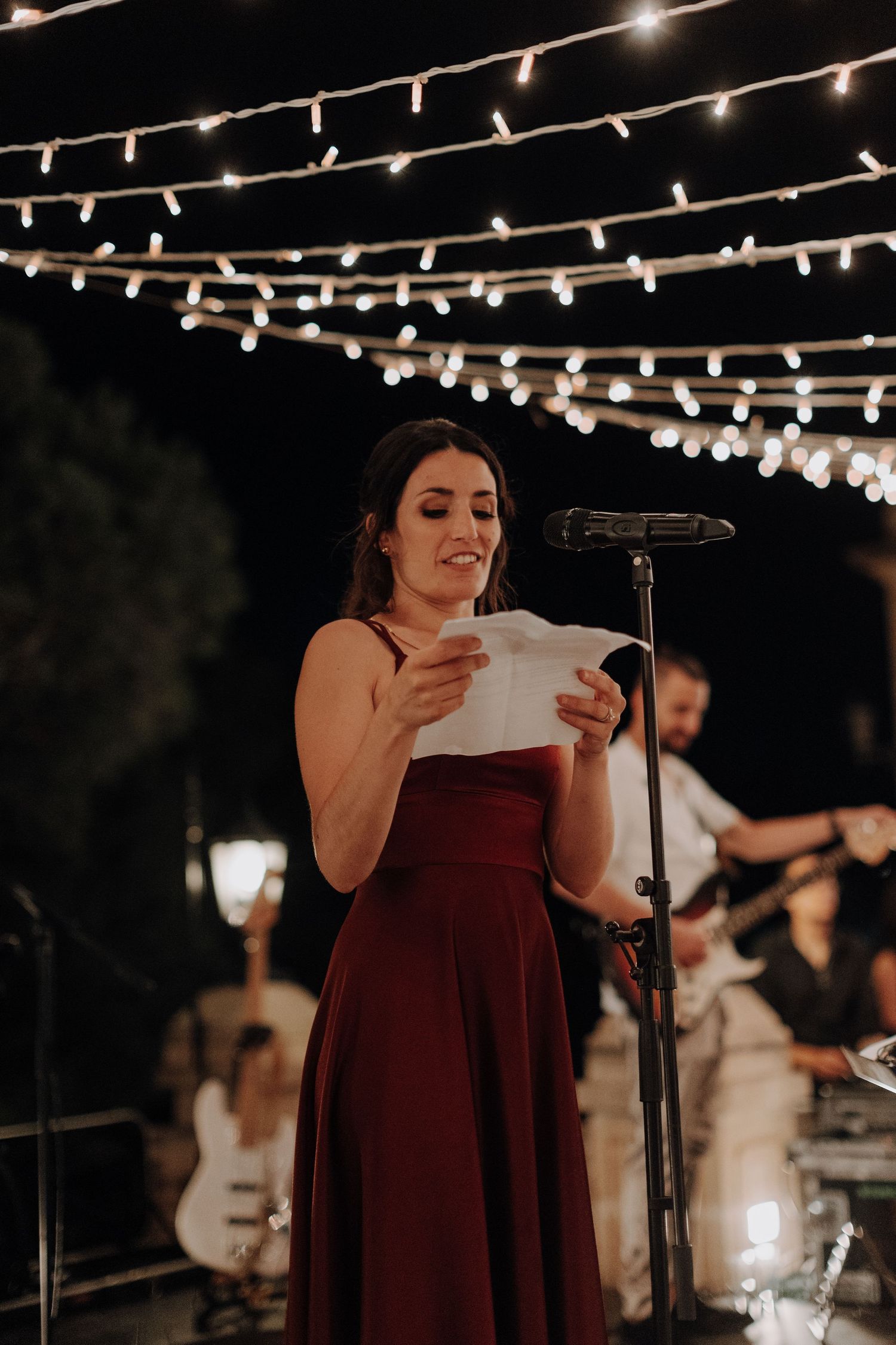 A woman in burgundy dress reads from a paper under string lights at an outdoor evening event.