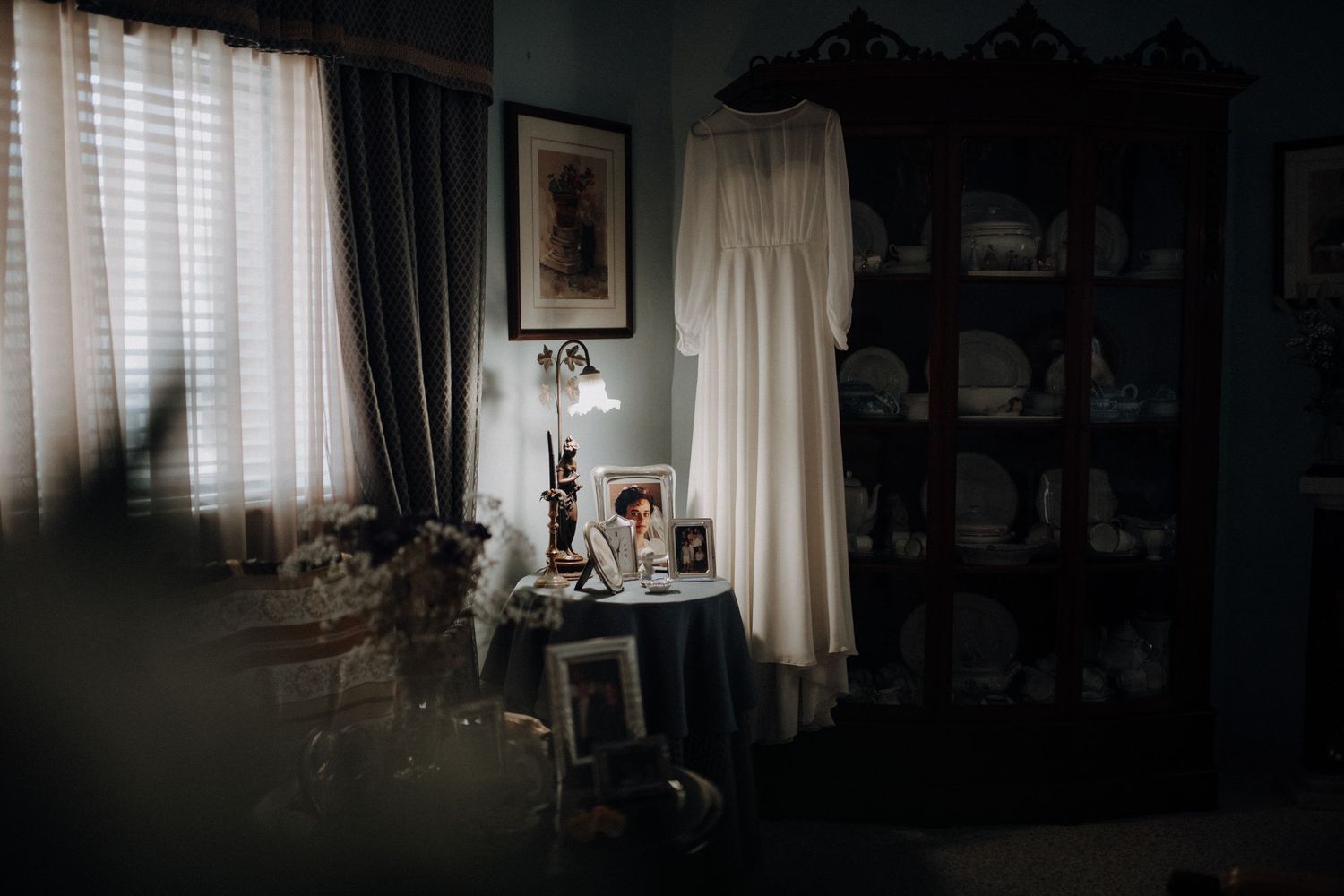 Moody interior shot of a wedding dress hanging in front of a window with vintage decor.