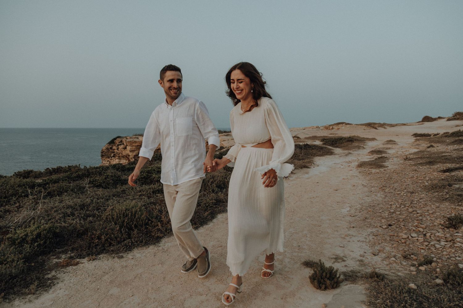 A couple in white outfits walks together along a coastal cliff path at sunset.