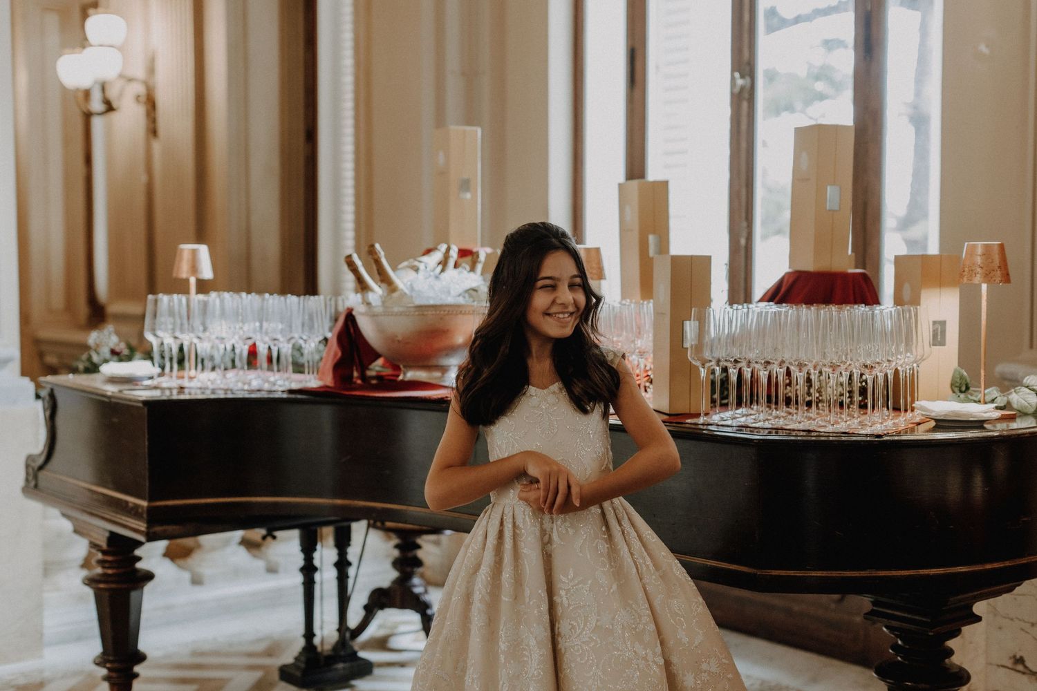 A bride in a ballgown poses beside a grand piano with champagne glasses.