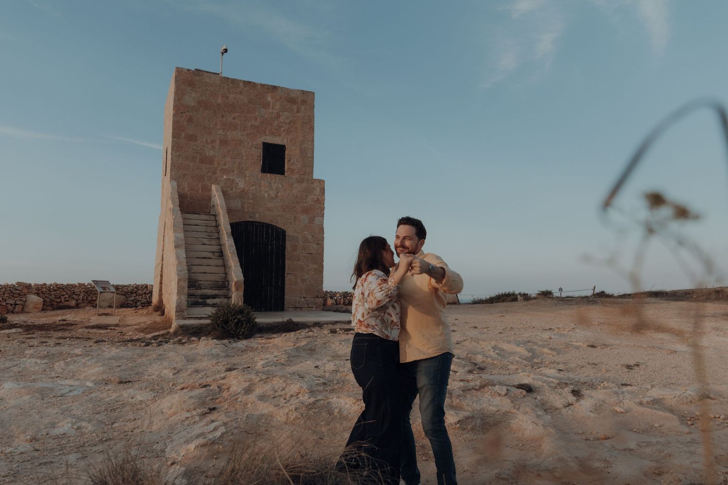 A couple embraces in a desert landscape with a rustic stone building in the background during sunset.