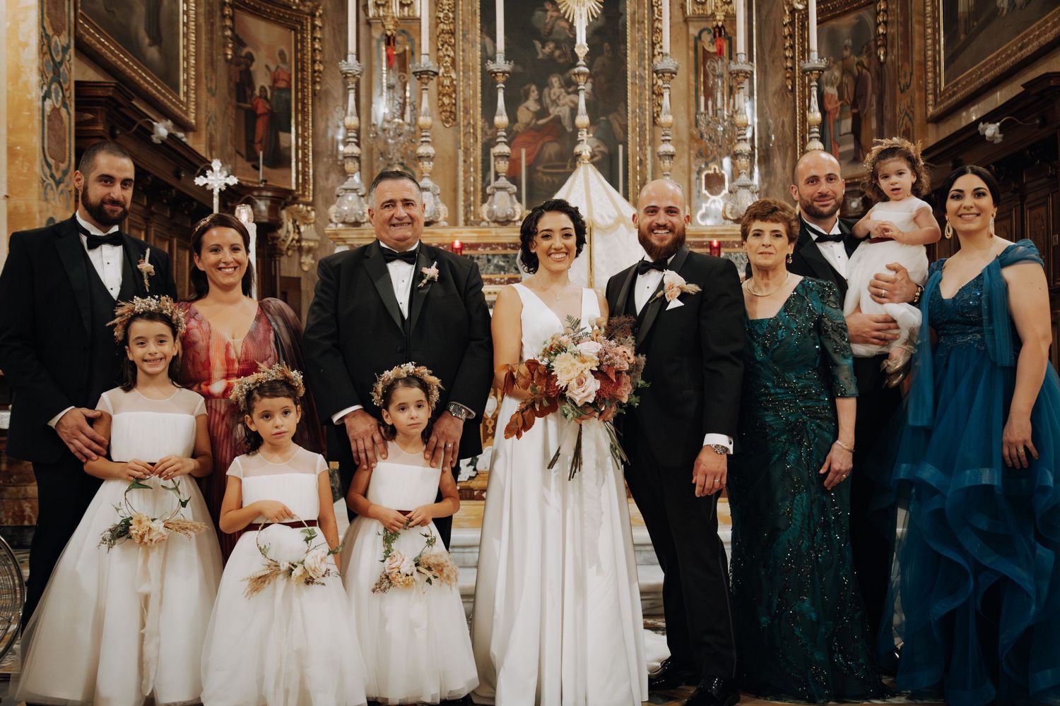 Extended wedding party poses formally in front of decorated church altar with flower girls in white dresses.