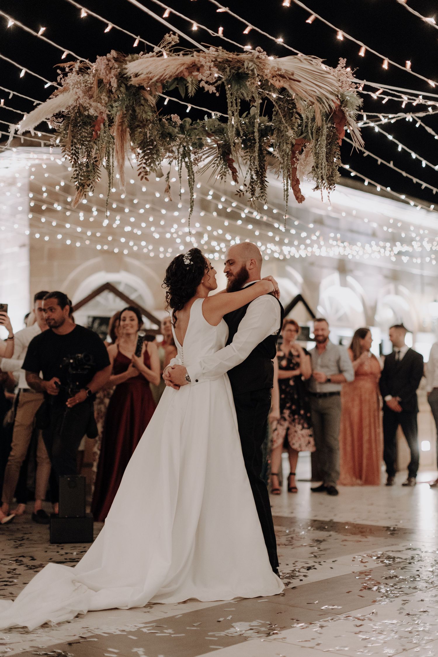Couple shares their first dance under romantic twinkle lights and floral arrangements at indoor wedding reception.