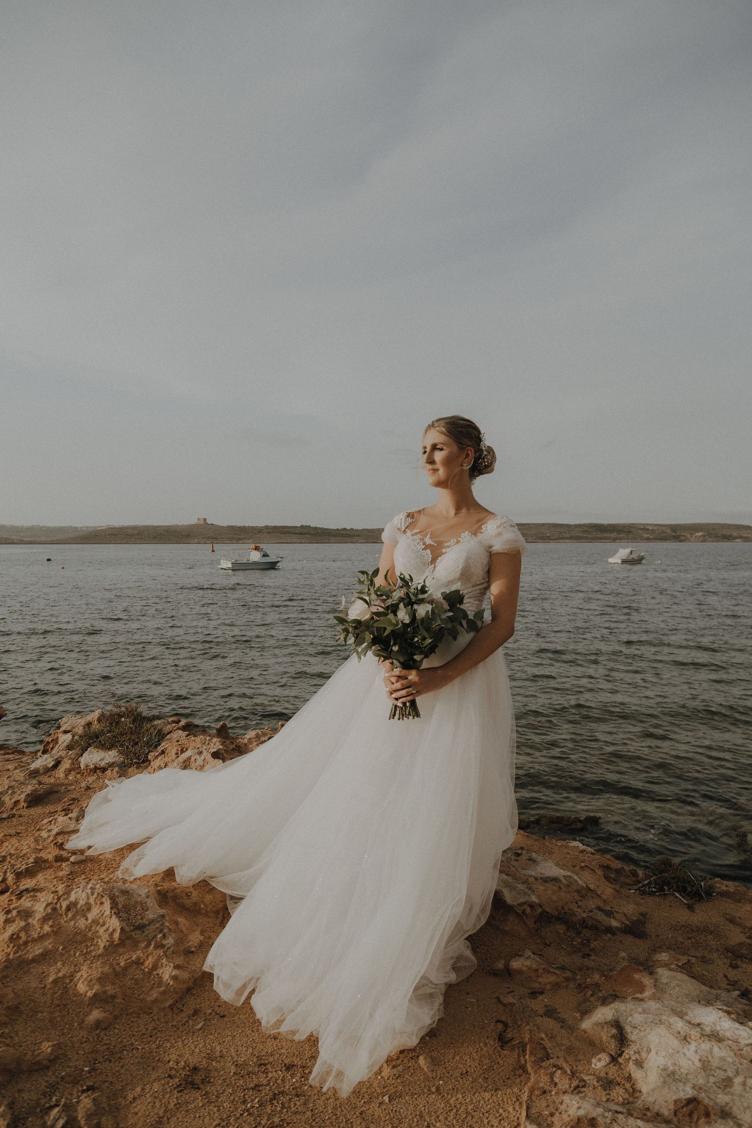 Elegant bride in flowing tulle gown poses on rocky coastline with boats in background.