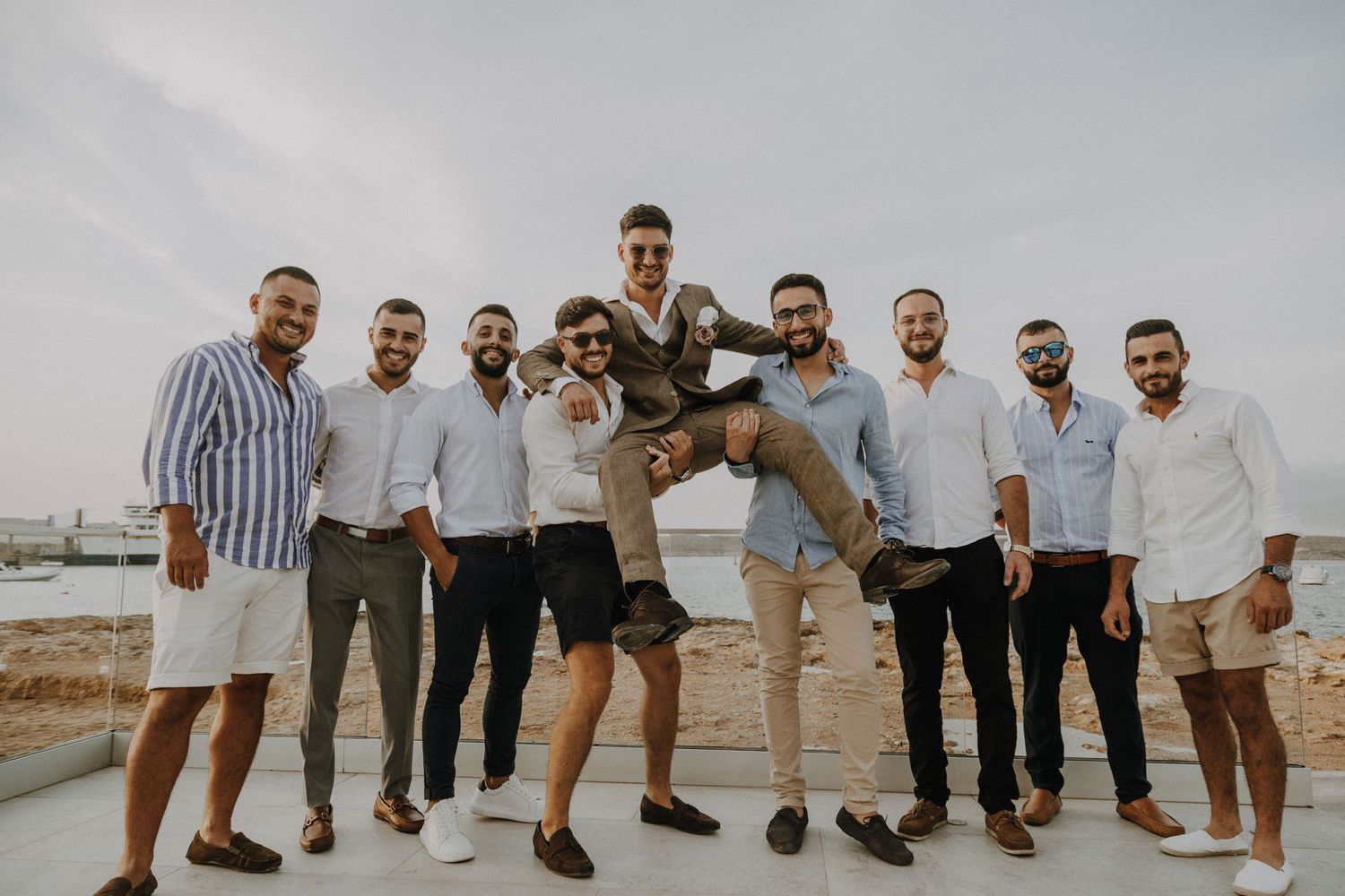 Groomsmen lift the groom in celebration at a waterfront wedding location.