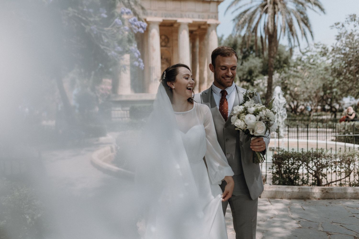 A wedding couple poses in front of a classical building with palm trees.