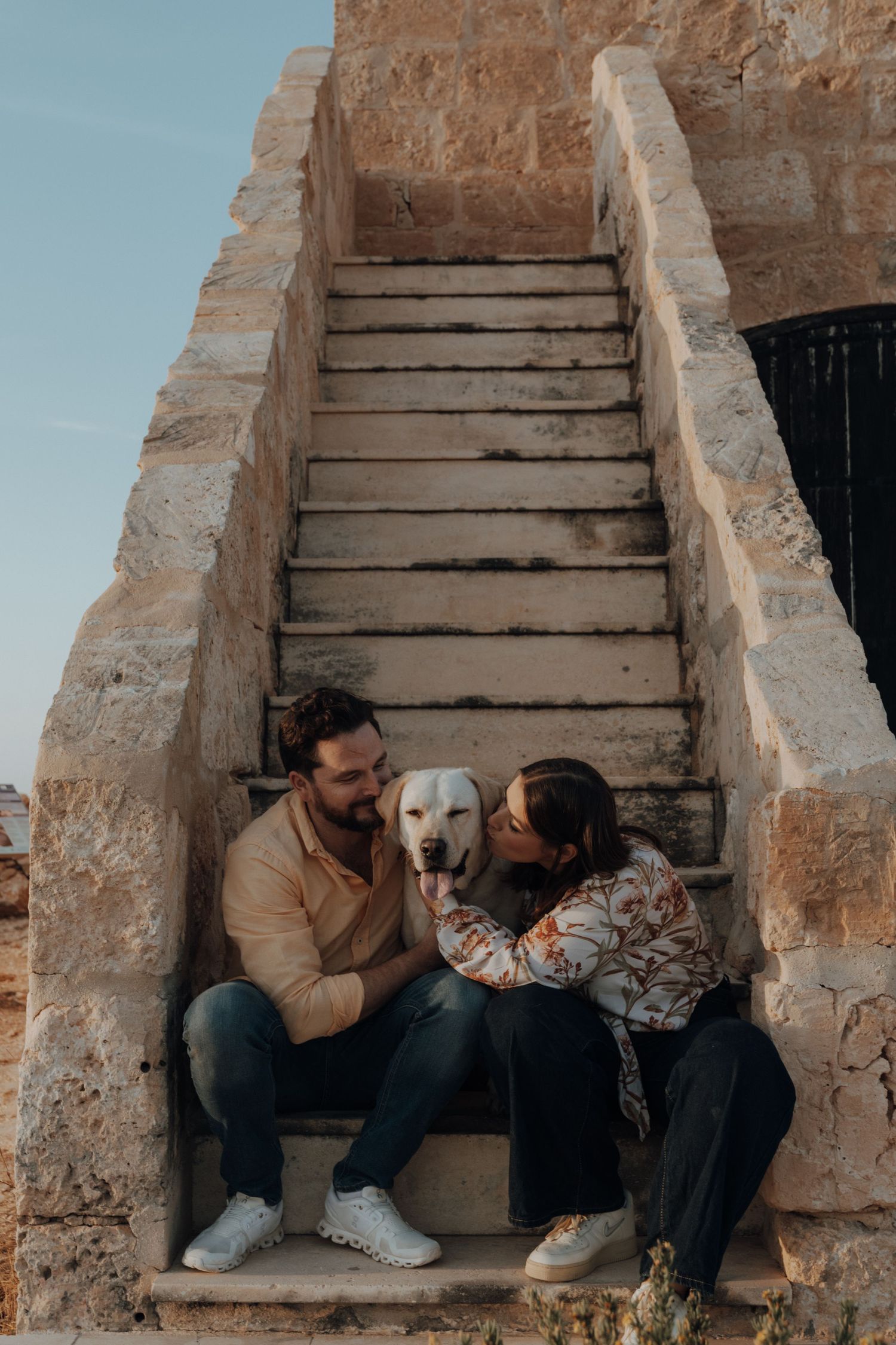 A couple sits intimately on stone steps against a rustic wall in warm lighting.