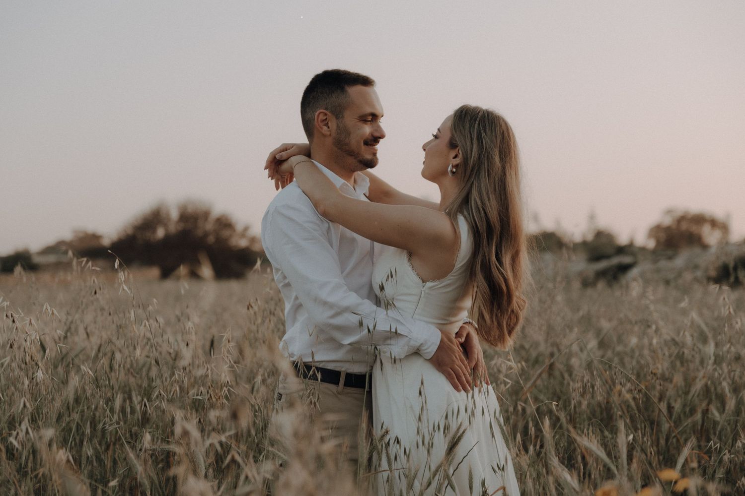 Couple embracing in golden field at sunset, woman in white dress with arms around man in white shirt.