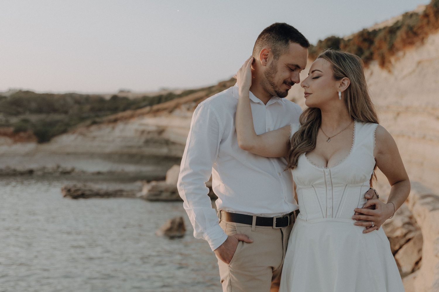 Couple in white outfits embracing on a rocky beach at golden hour during an engagement photoshoot.