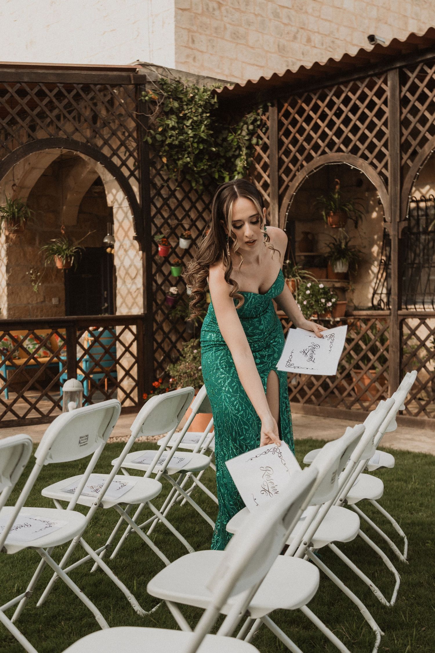 Person in emerald green dress placing items on white folding chairs at outdoor venue.