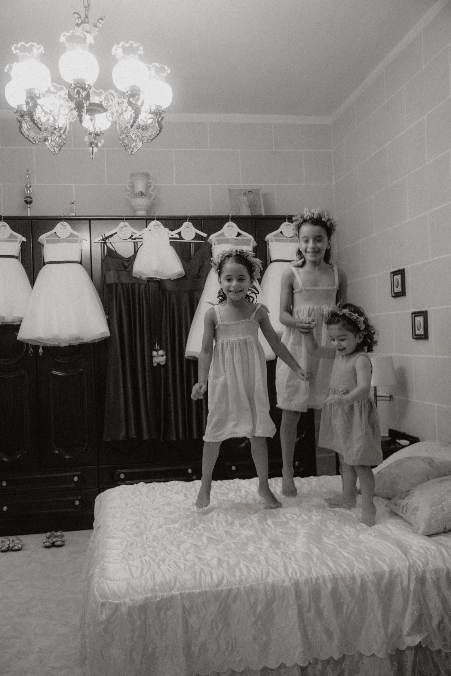 Children jumping and playing on a large white bed with flower girl dresses hanging above.