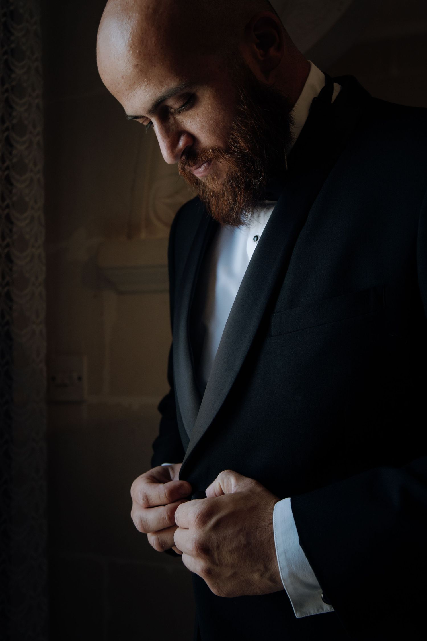 Groom in dark tuxedo adjusts cufflinks in moody lighting.