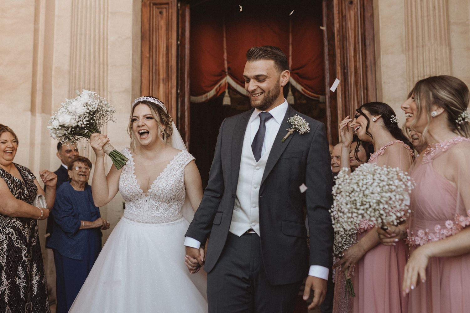 Newlyweds exit church surrounded by wedding party in pink dresses.